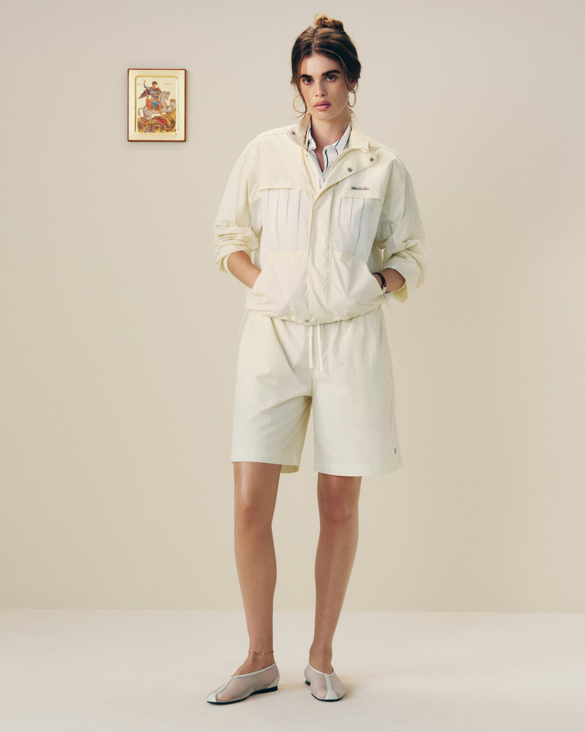 A woman in a Nylon Track Jacket and shorts stands indoors with hands in pockets near a framed wall picture.