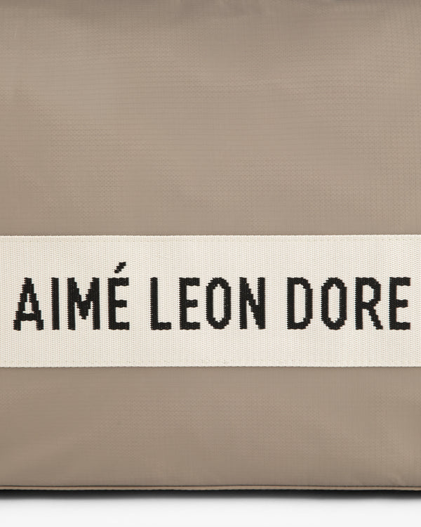 A beige nylon duffle bag with a white band featuring AIMÉ LEON DORE in black capital letters.