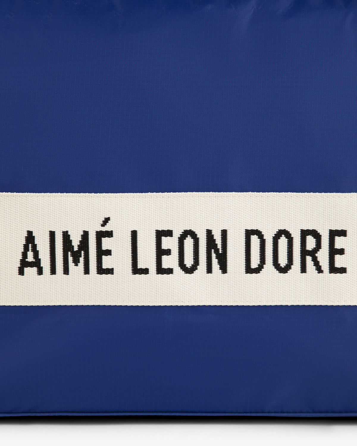 A blue nylon duffle bag with a white strap and black AIMÉ LEON DORE text.