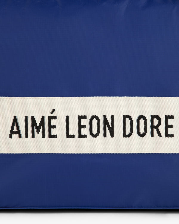 A blue nylon duffle bag with a white strap and black AIMÉ LEON DORE text.