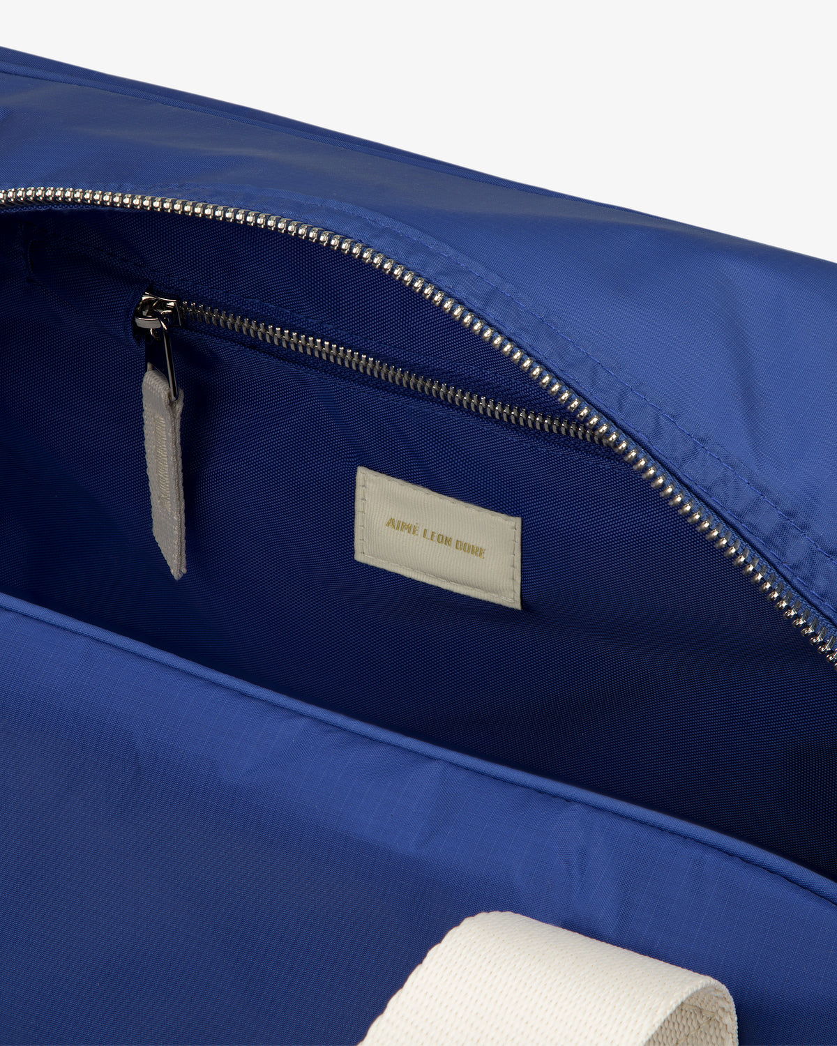 Close-up of an open Nylon Duffle Bag with a zippered pocket, white label inside, and part of a white strap visible.