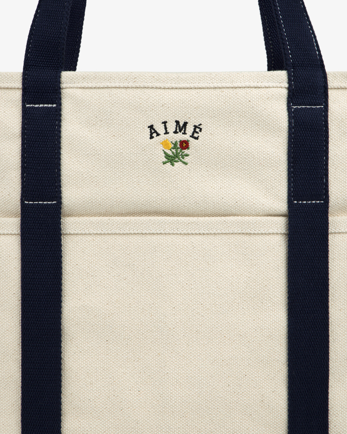 Close-up of the Large Crest Contrast Tote Bag with navy handles and AIMÉ embroidered above a small yellow-red flower.