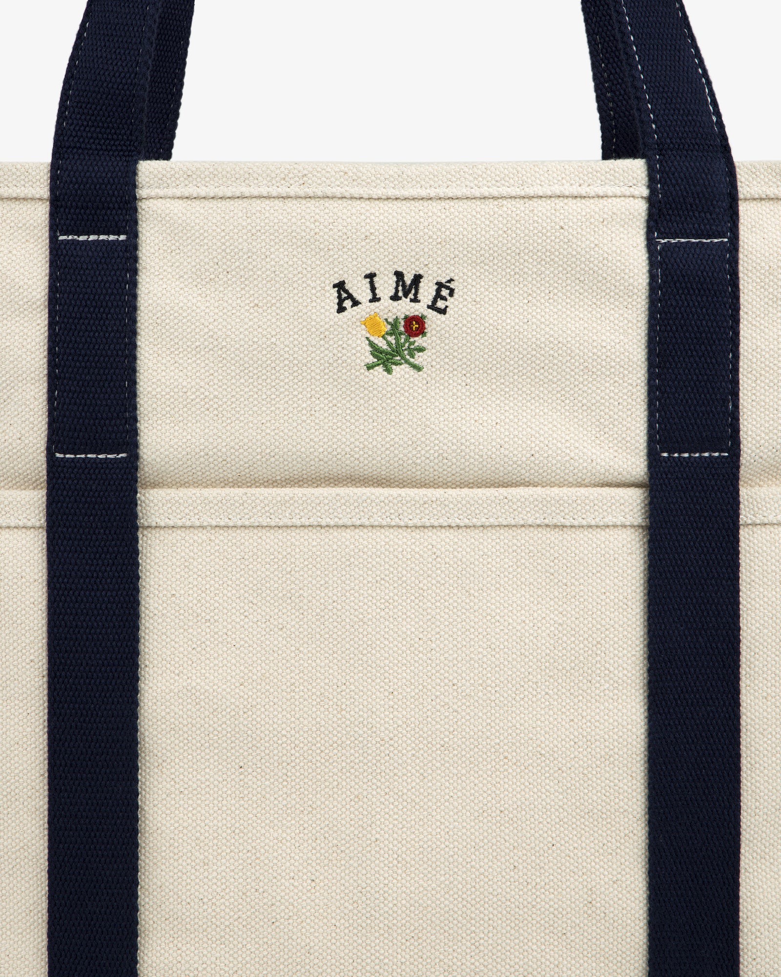 Close-up of the Large Crest Contrast Tote Bag with navy handles and AIMÉ embroidered above a small yellow-red flower.
