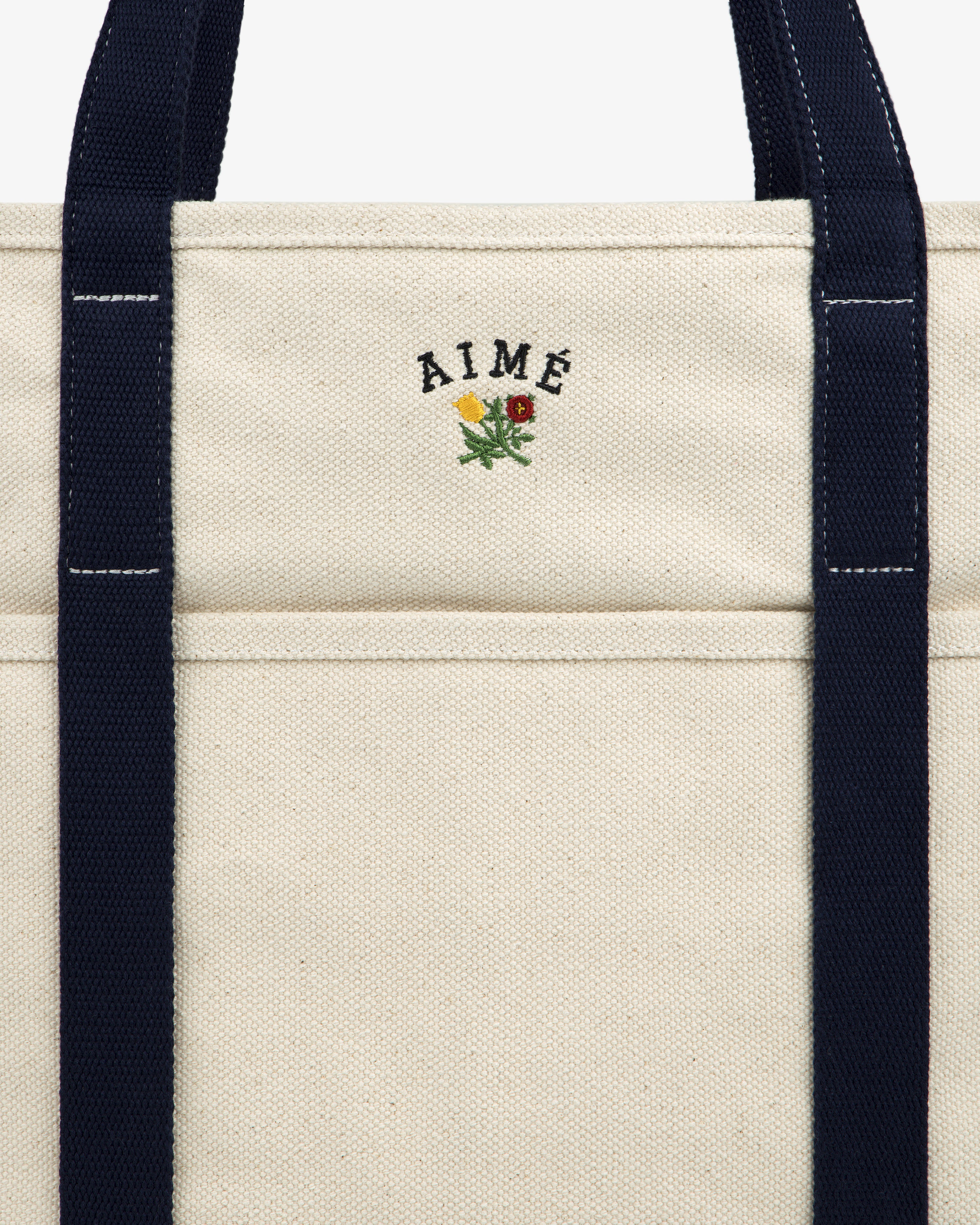 Close-up of the Large Crest Contrast Tote Bag with navy handles and AIMÉ embroidered above a small yellow-red flower.