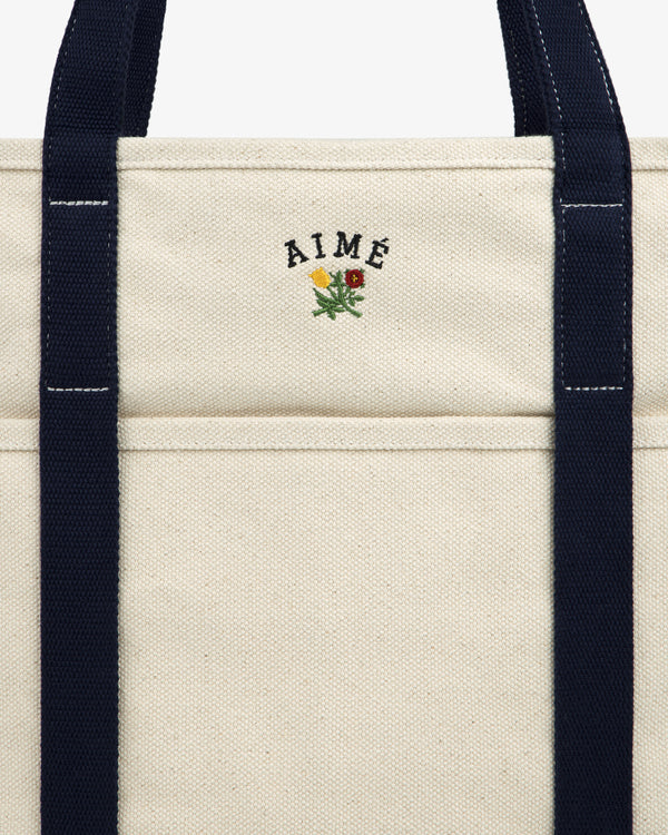 Close-up of the Large Crest Contrast Tote Bag with navy handles and AIMÉ embroidered above a small yellow-red flower.