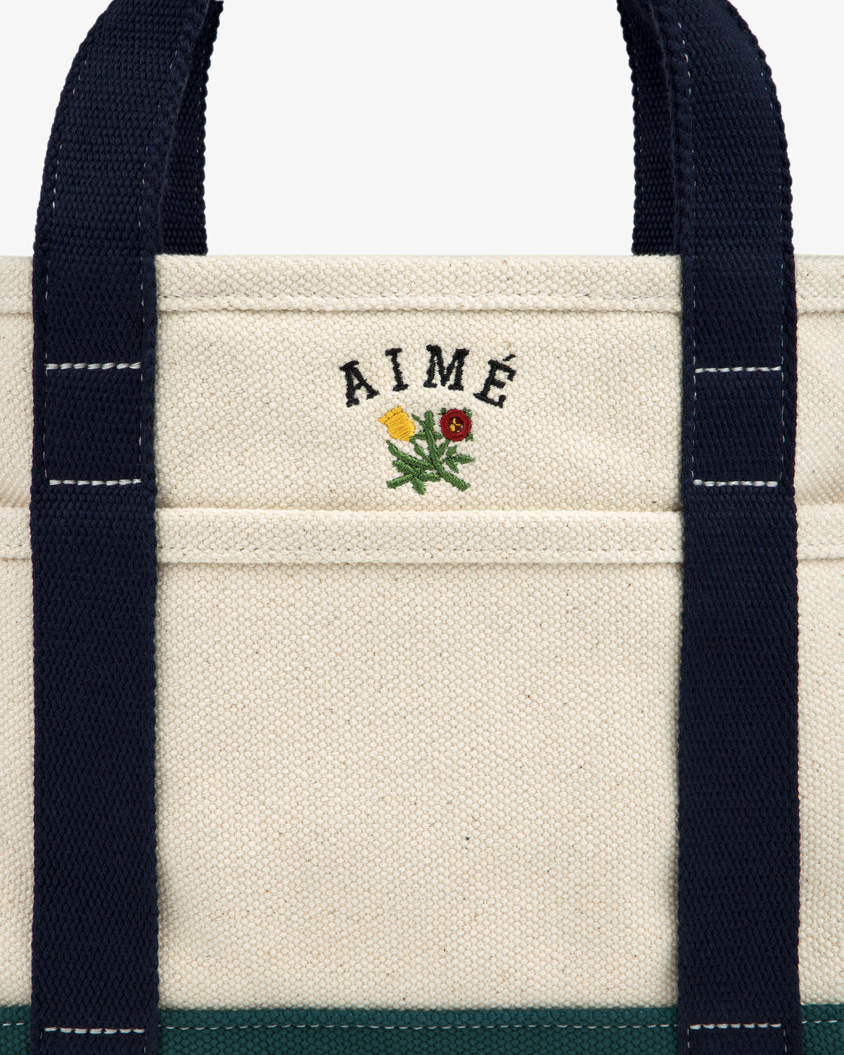 Mini Crest Contrast Tote Bag: cream canvas with navy handles, AIMÉ text, and yellow-red flower above the pocket.