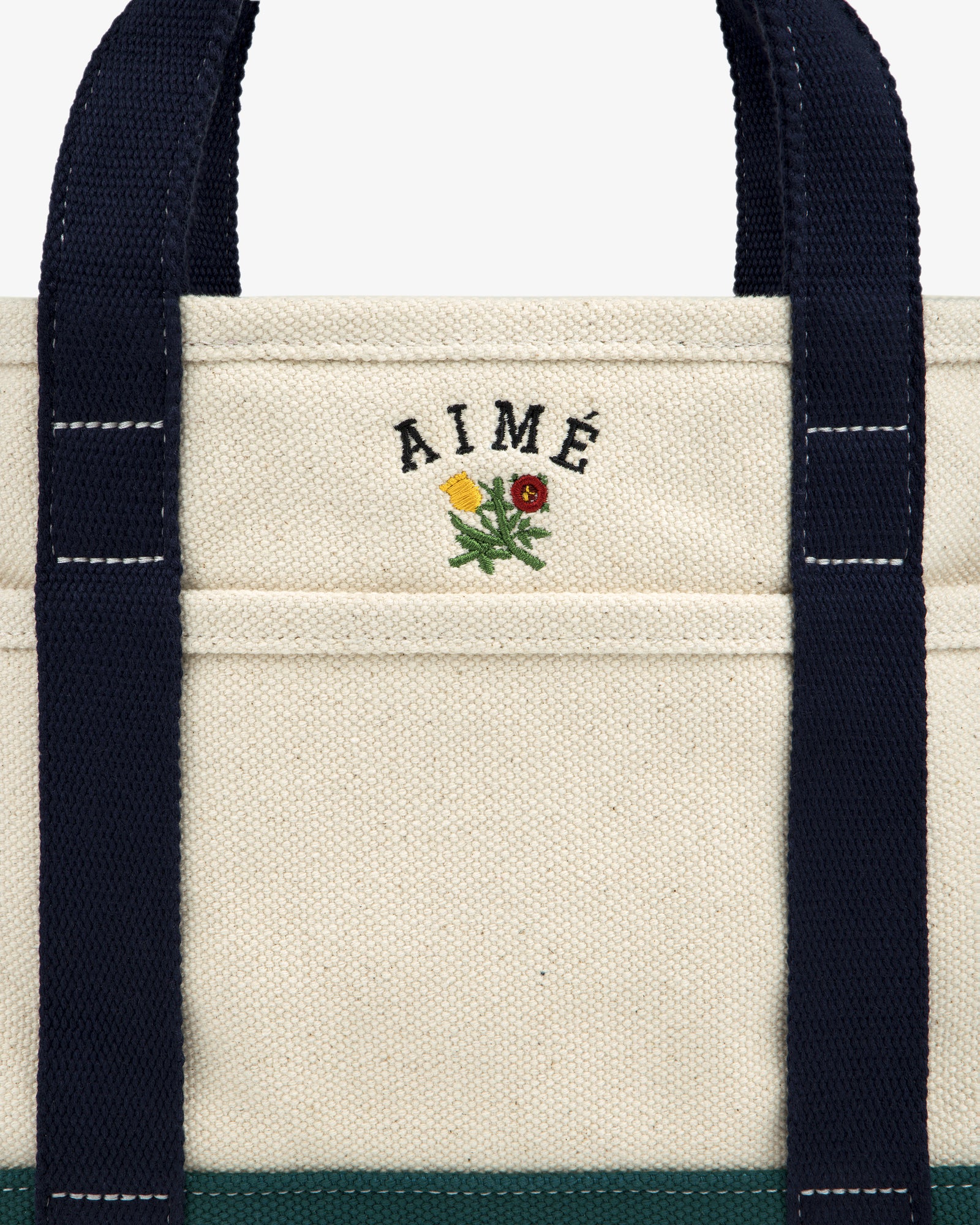 Mini Crest Contrast Tote Bag: cream canvas with navy handles, AIMÉ text, and yellow-red flower above the pocket.