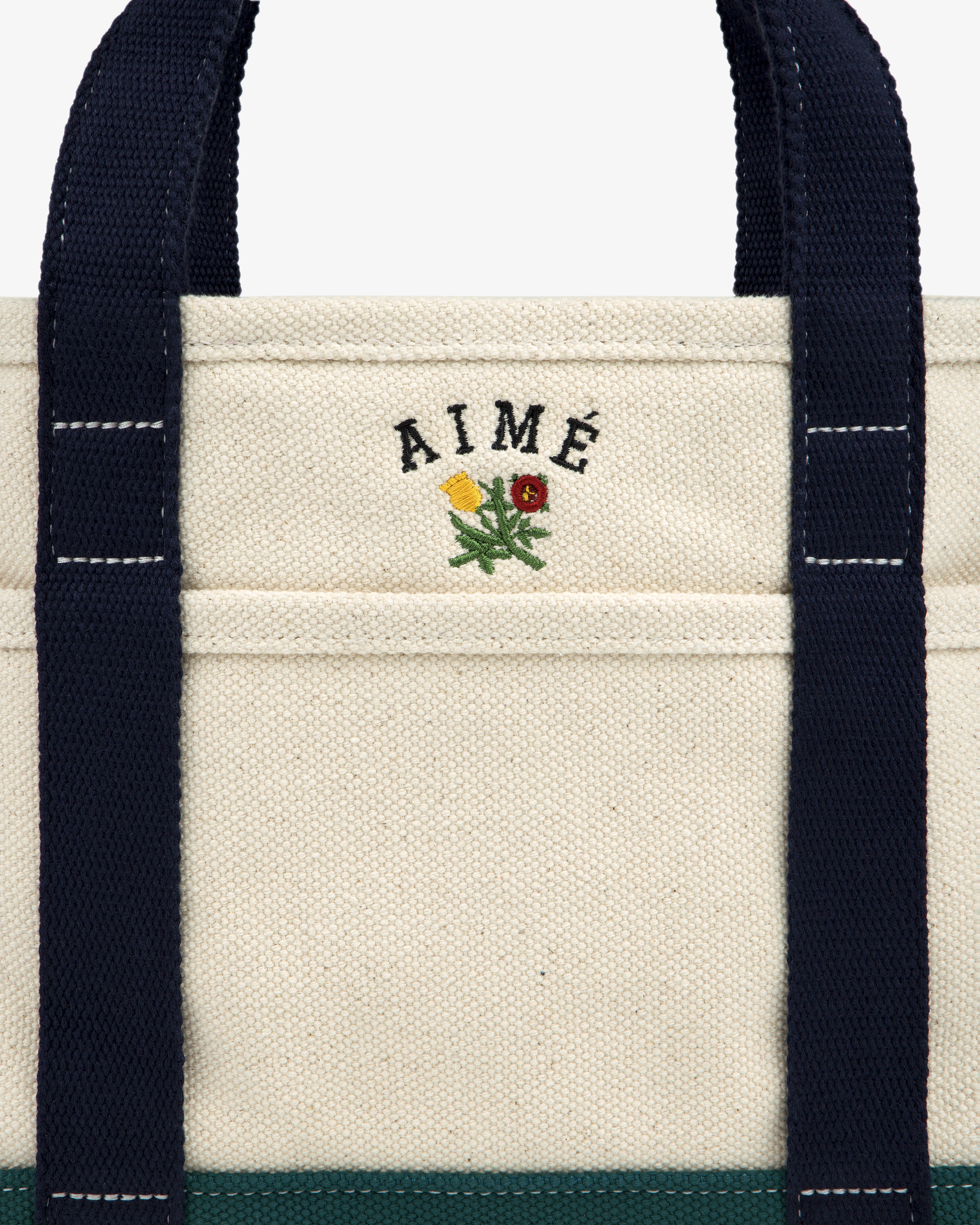 Mini Crest Contrast Tote Bag: cream canvas with navy handles, AIMÉ text, and yellow-red flower above the pocket.