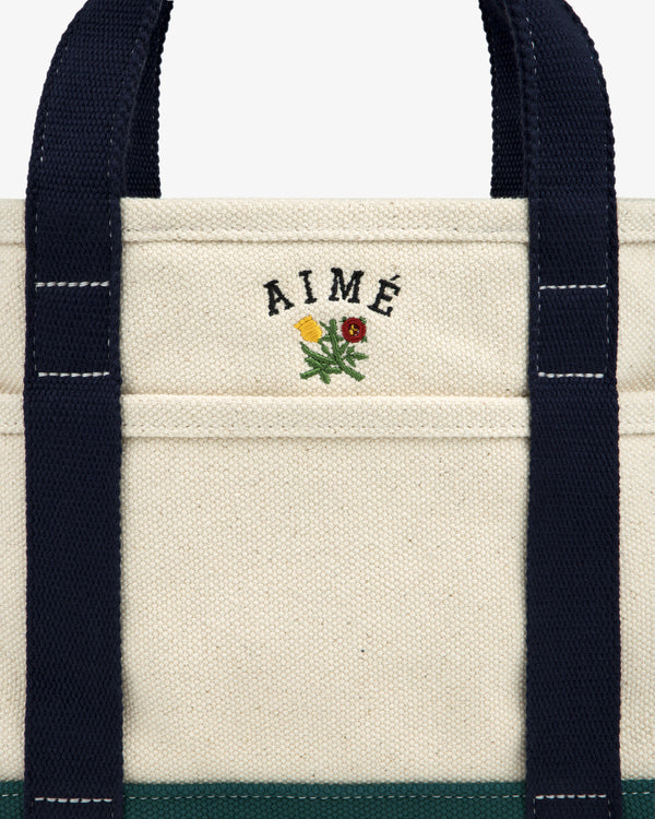 Mini Crest Contrast Tote Bag: cream canvas with navy handles, AIMÉ text, and yellow-red flower above the pocket.