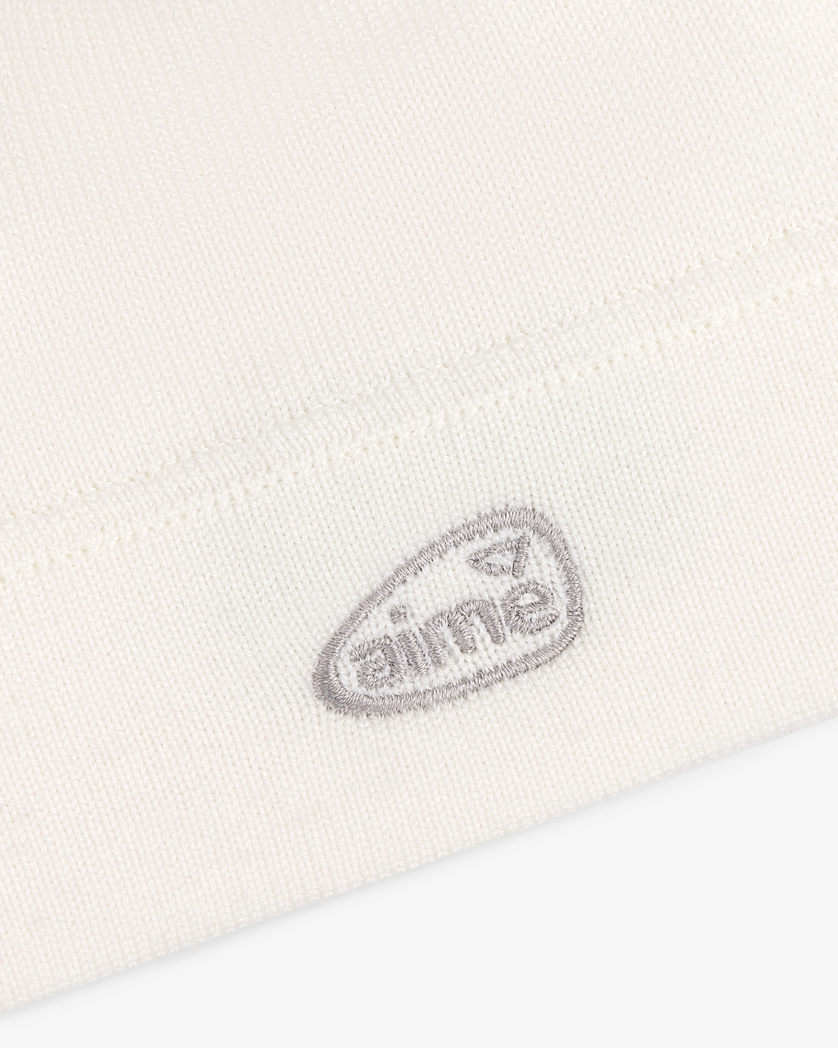 Close-up of the ALD Golf Skull Cap's white knit fabric with a small gray aimé logo embroidered near the hem.