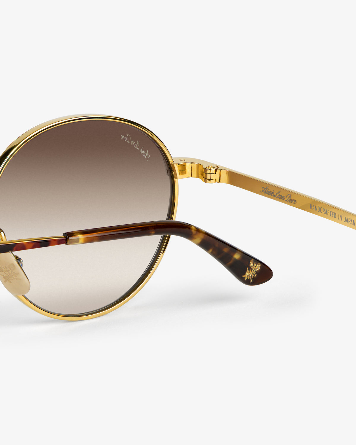 Close-up of Giórgios Sunglasses: gold and tortoiseshell, round lenses, HANDCRAFTED IN JAPAN engraved on the arm.