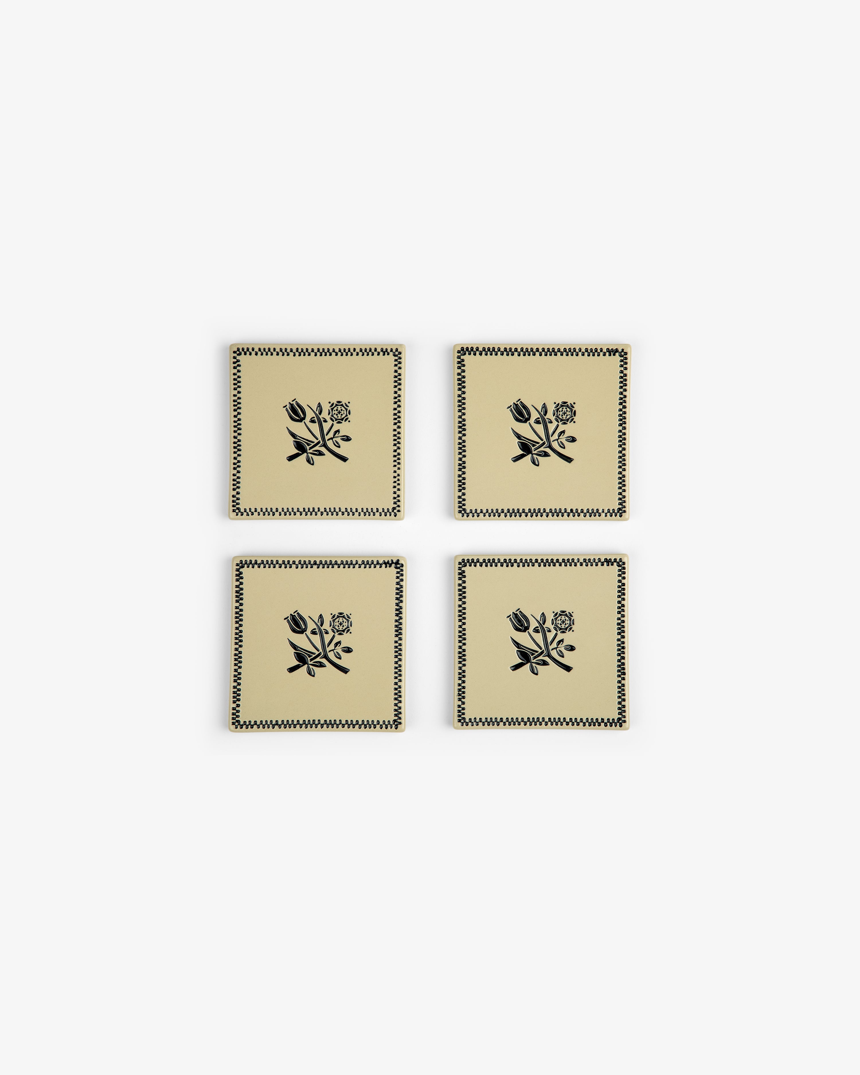 The Crest Coaster Set (4 pcs) features beige coasters with black floral designs and zigzag borders, arranged in a grid.