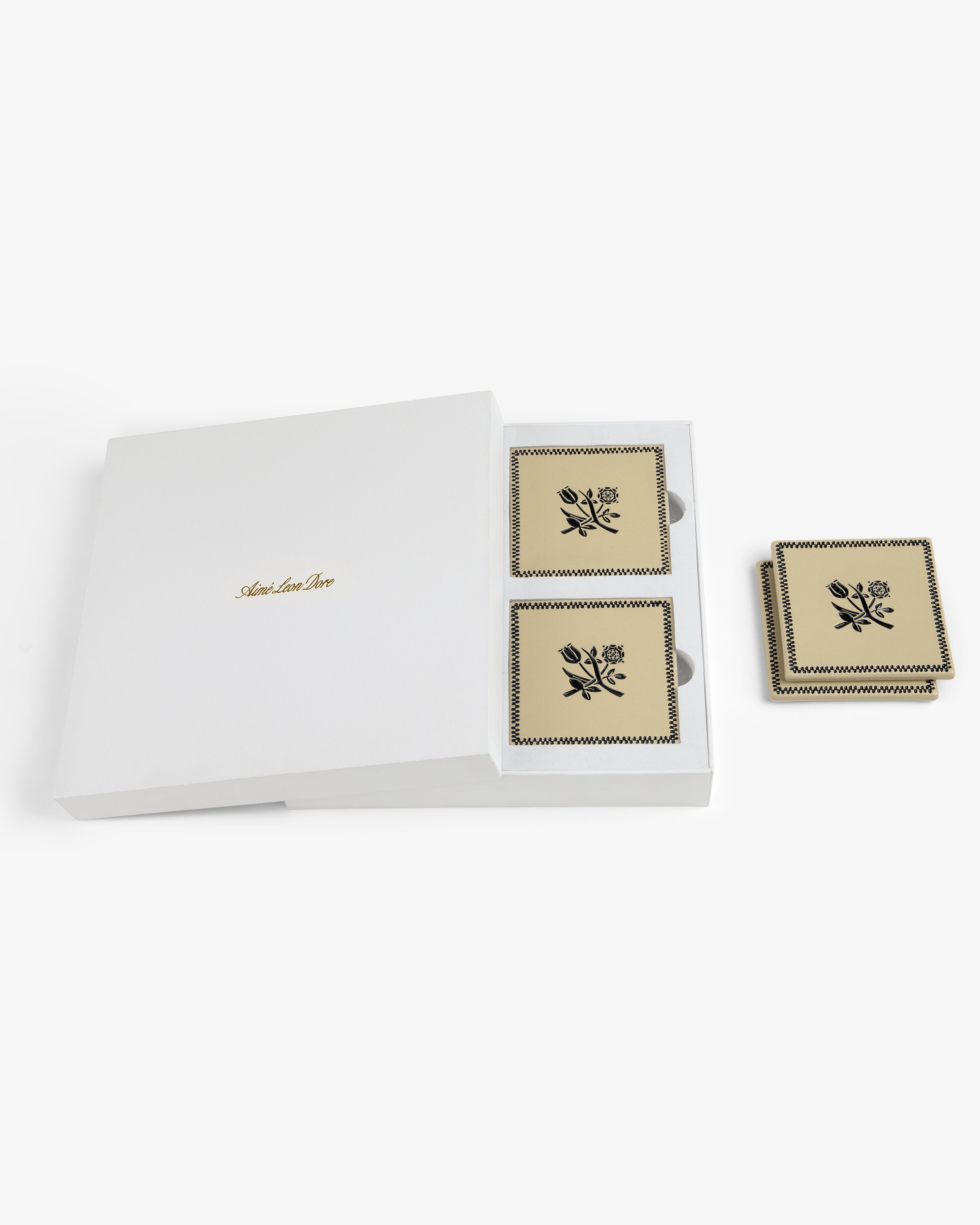 The Crest Coaster (Set of 4) comes in a white box with gold text, featuring square beige coasters with black florals.