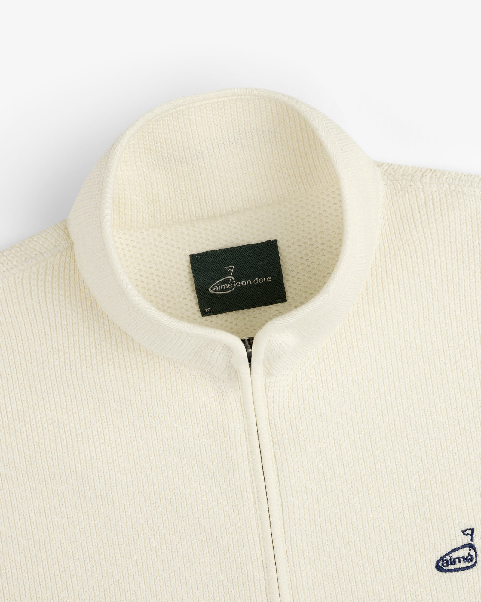 Close-up of the cream ALD Golf Piqué Full Zip Sweater with textured fabric, stand collar, and inner Aimé Leon Dore label.