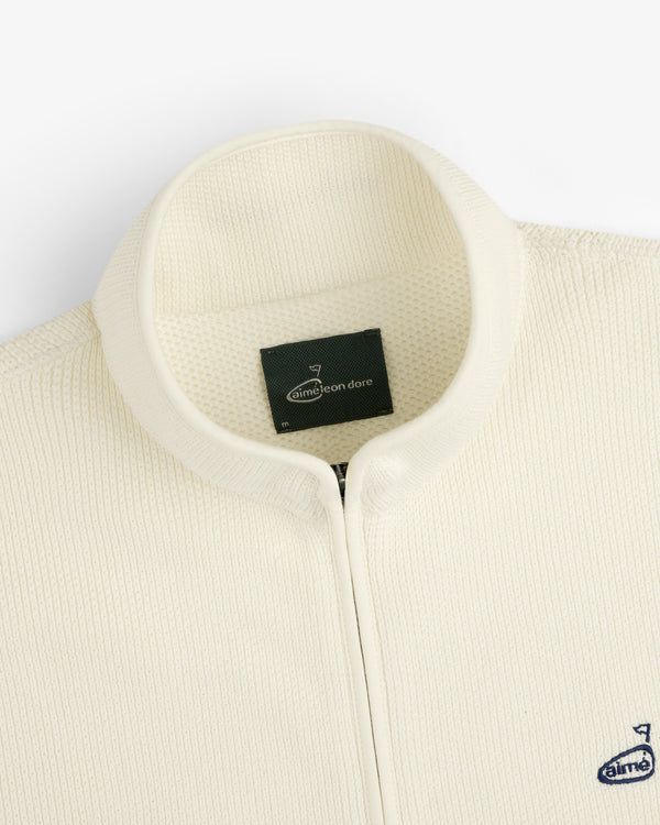 Close-up of the cream ALD Golf Piqué Full Zip Sweater with textured fabric, stand collar, and inner Aimé Leon Dore label.