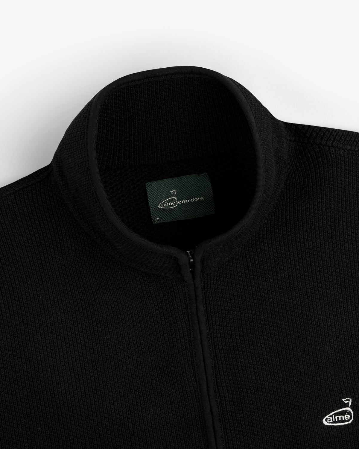 Close-up of an ALD Golf Piqué Full Zip Sweater in black with label and small embroidered chest logo.