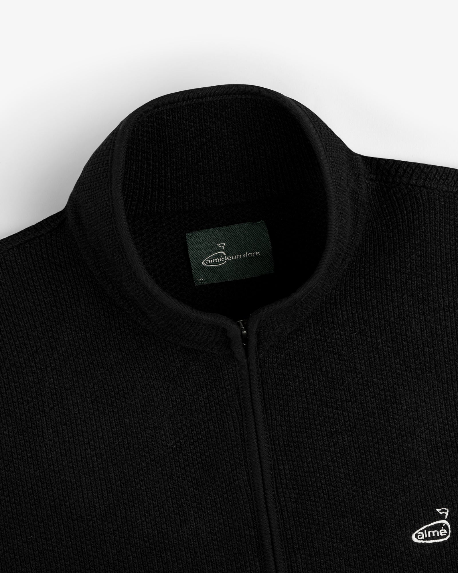 Close-up of an ALD Golf Piqué Full Zip Sweater in black with label and small embroidered chest logo.