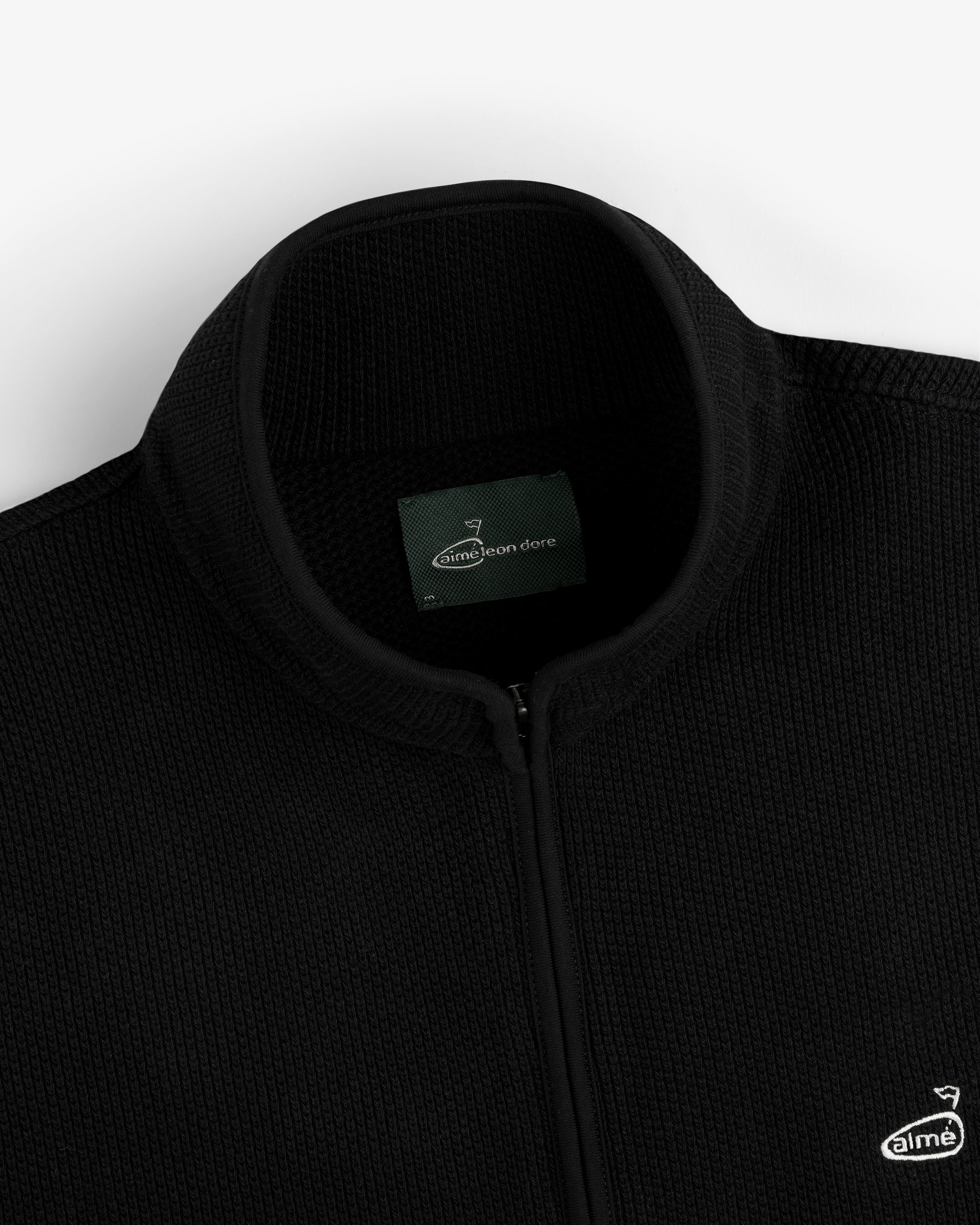 Close-up of an ALD Golf Piqué Full Zip Sweater in black with label and small embroidered chest logo.