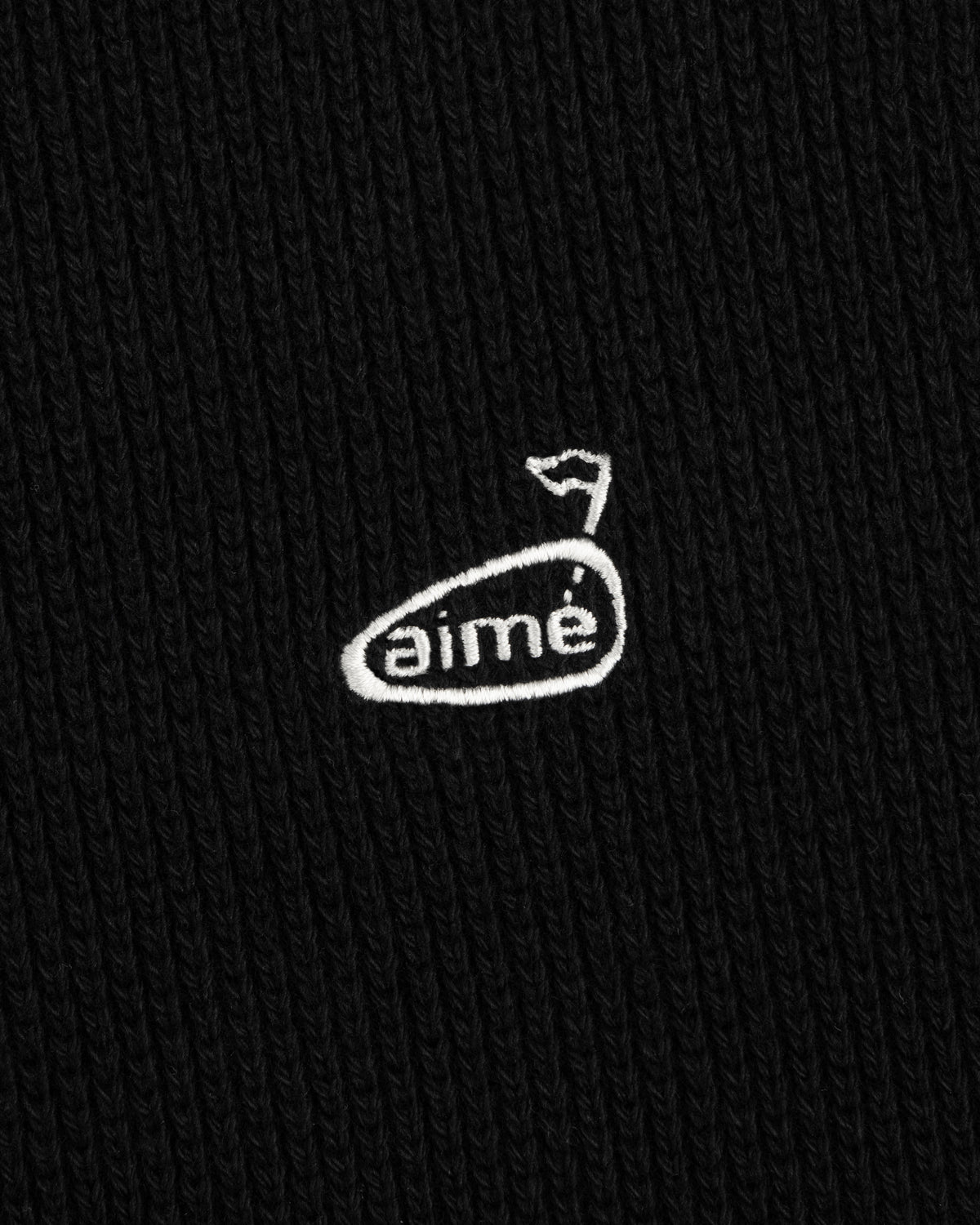 Close-up of the ALD Golf Piqué Full Zip Sweater’s black ribbed fabric with white aimé logo and a small flag above.