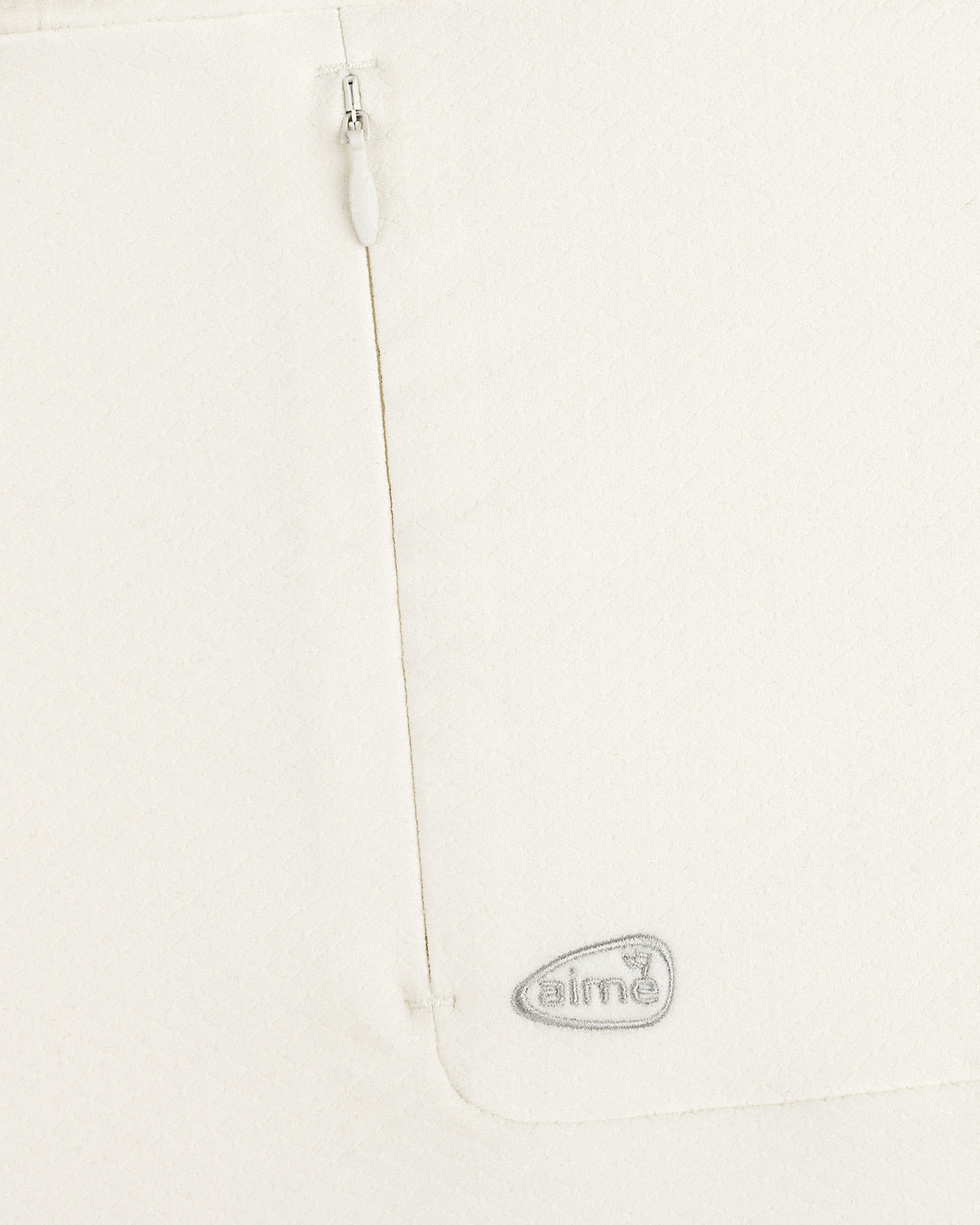 Close-up of the ALD Golf Quarter Zip Pullover’s white textured fabric with a zipper and gray “aimé” logo near the seam.