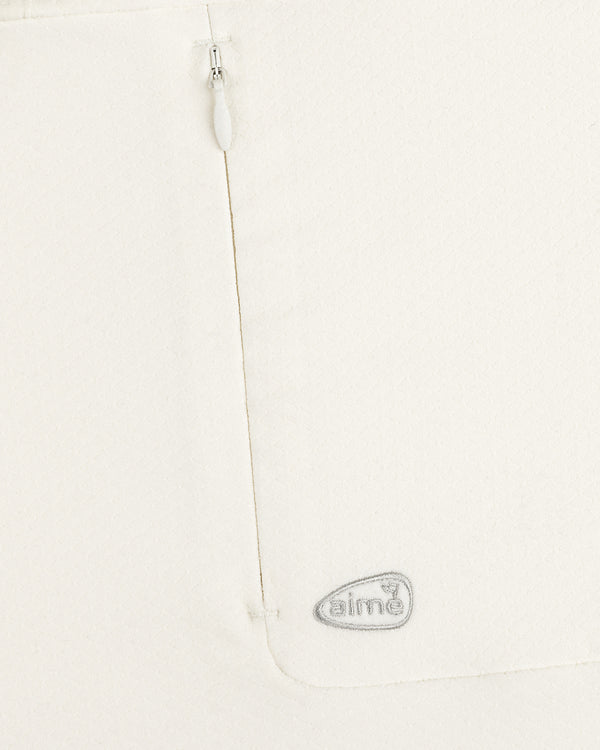 Close-up of the ALD Golf Quarter Zip Pullover’s white textured fabric with a zipper and gray “aimé” logo near the seam.