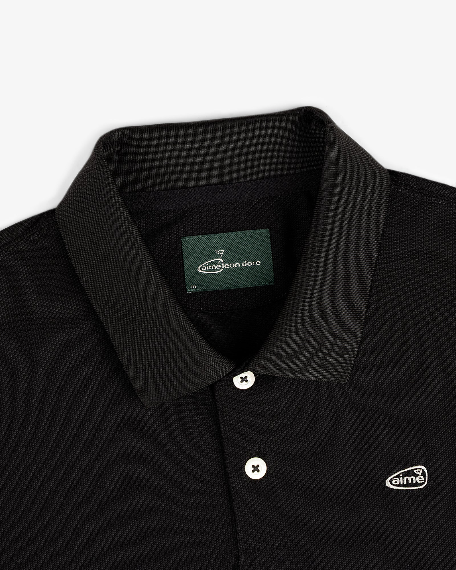 Close-up of the ALD Golf Polo, a black collared shirt with white buttons and a small Aimé Leon Dore chest logo patch.