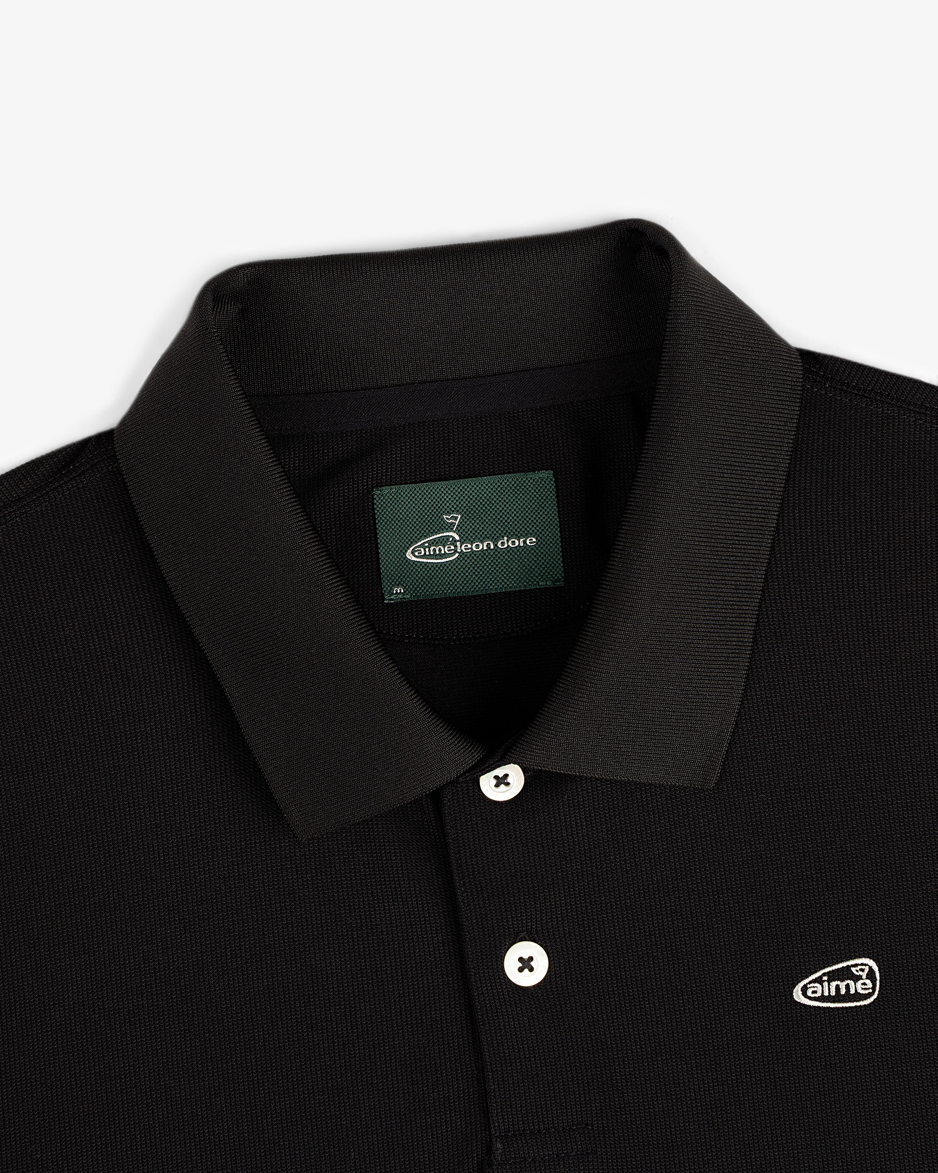 Close-up of the ALD Golf Polo, a black collared shirt with white buttons and a small Aimé Leon Dore chest logo patch.