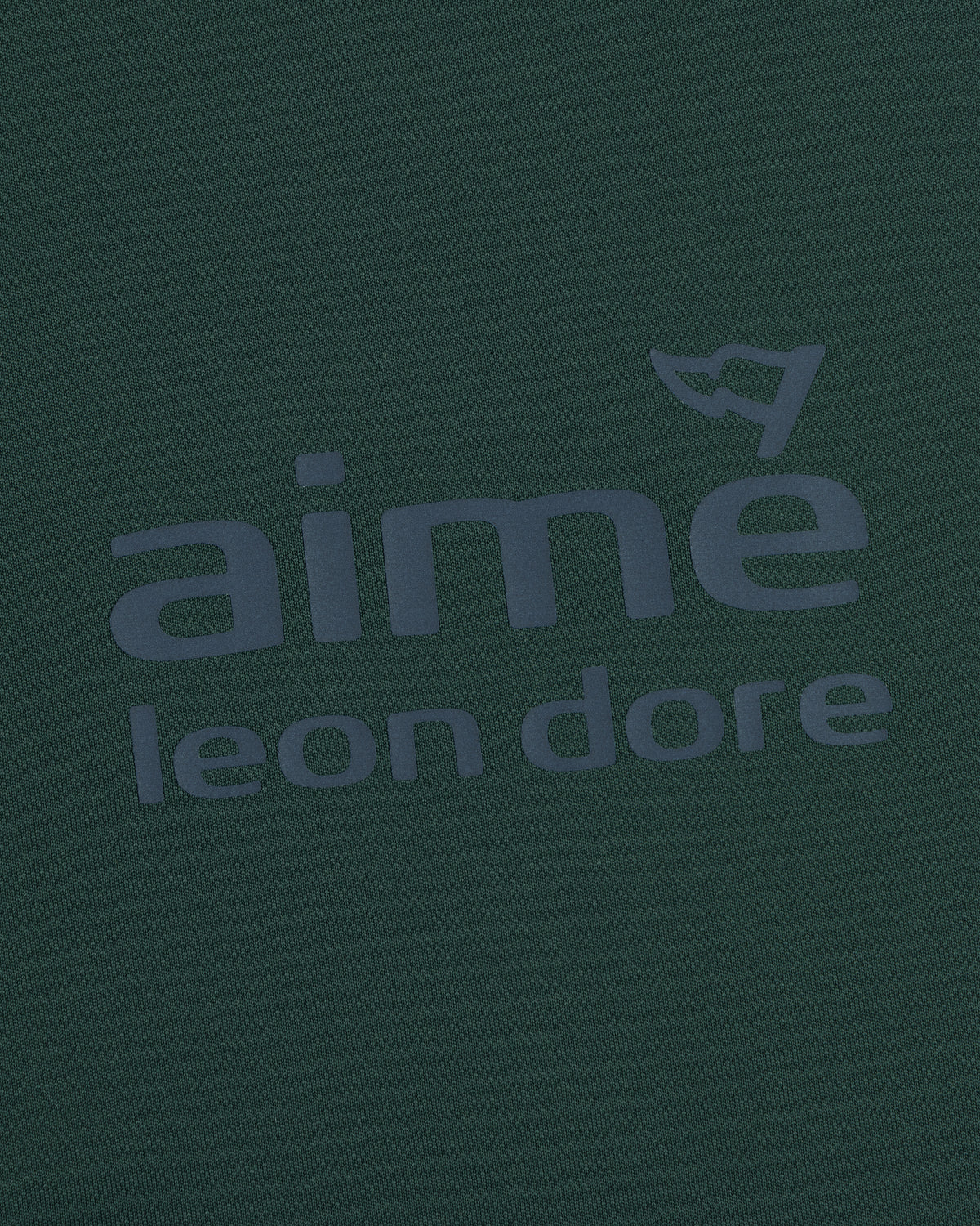 Close-up of the ALD Golf Tour Polo’s dark green fabric with bold aimé leon dore text and a flag graphic above the "e".