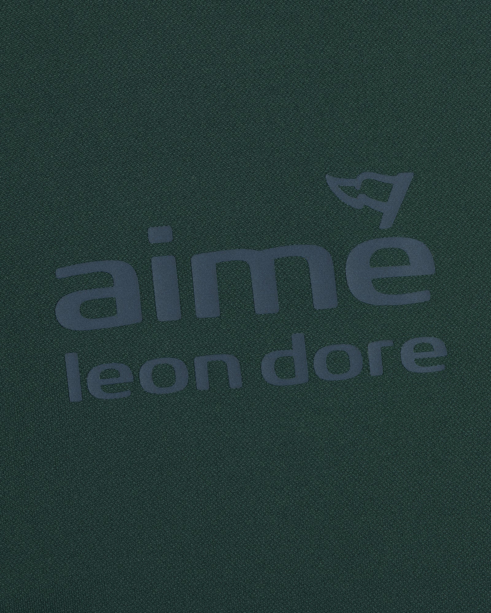 Close-up of the ALD Golf Tour Polo’s dark green fabric with bold aimé leon dore text and a flag graphic above the "e".