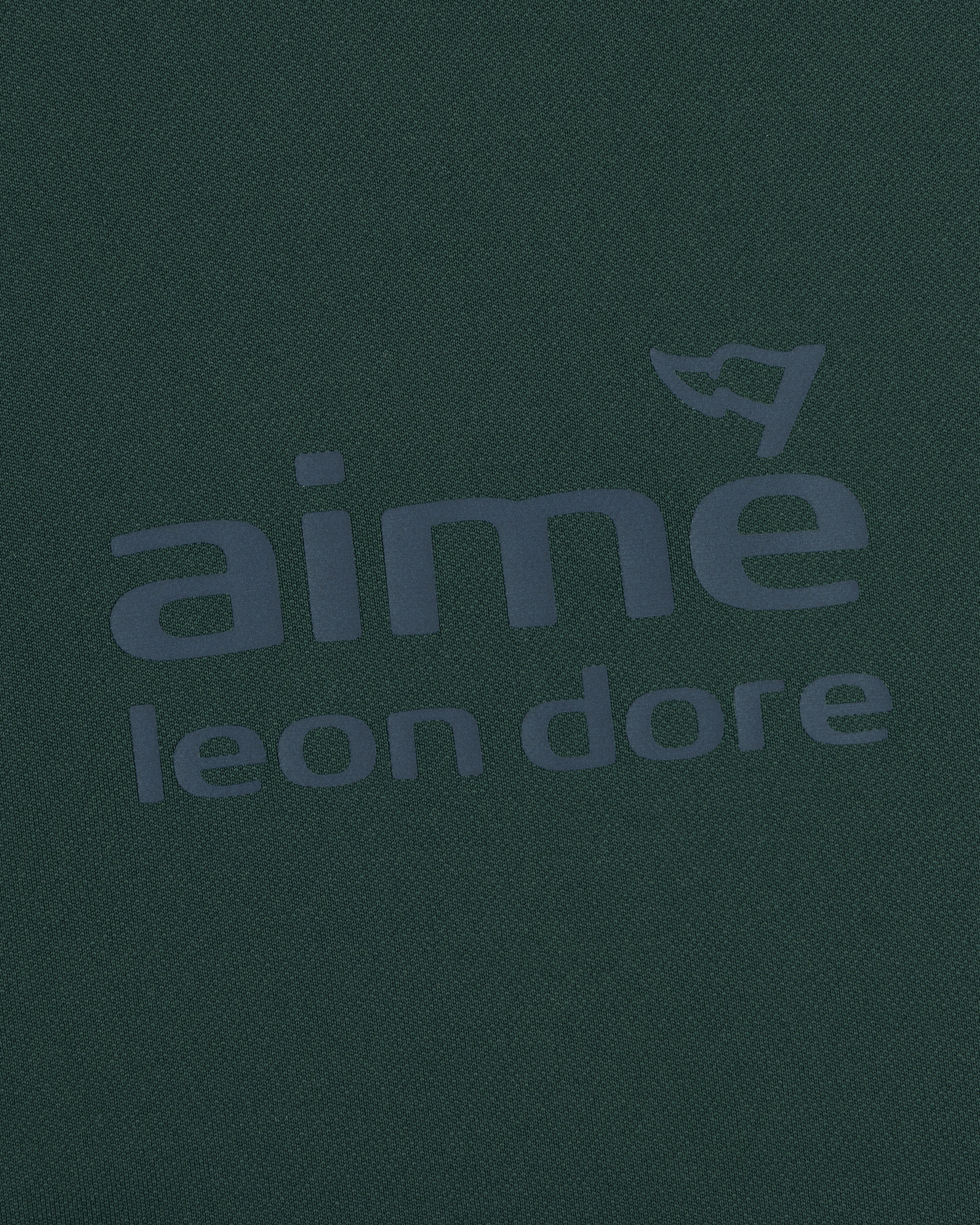 Close-up of the ALD Golf Tour Polo’s dark green fabric with bold aimé leon dore text and a flag graphic above the "e".