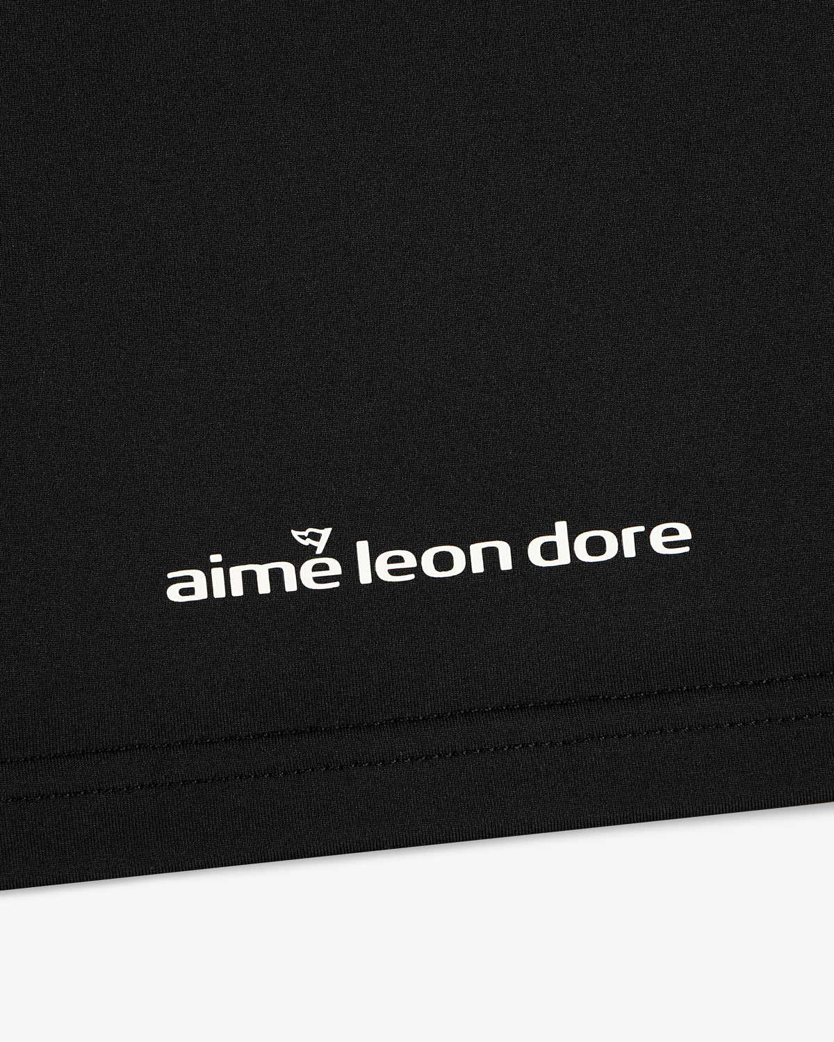 Close-up of black ALD Golf Performance Tee with white aime leon dore logo and stitching near the bottom edge.