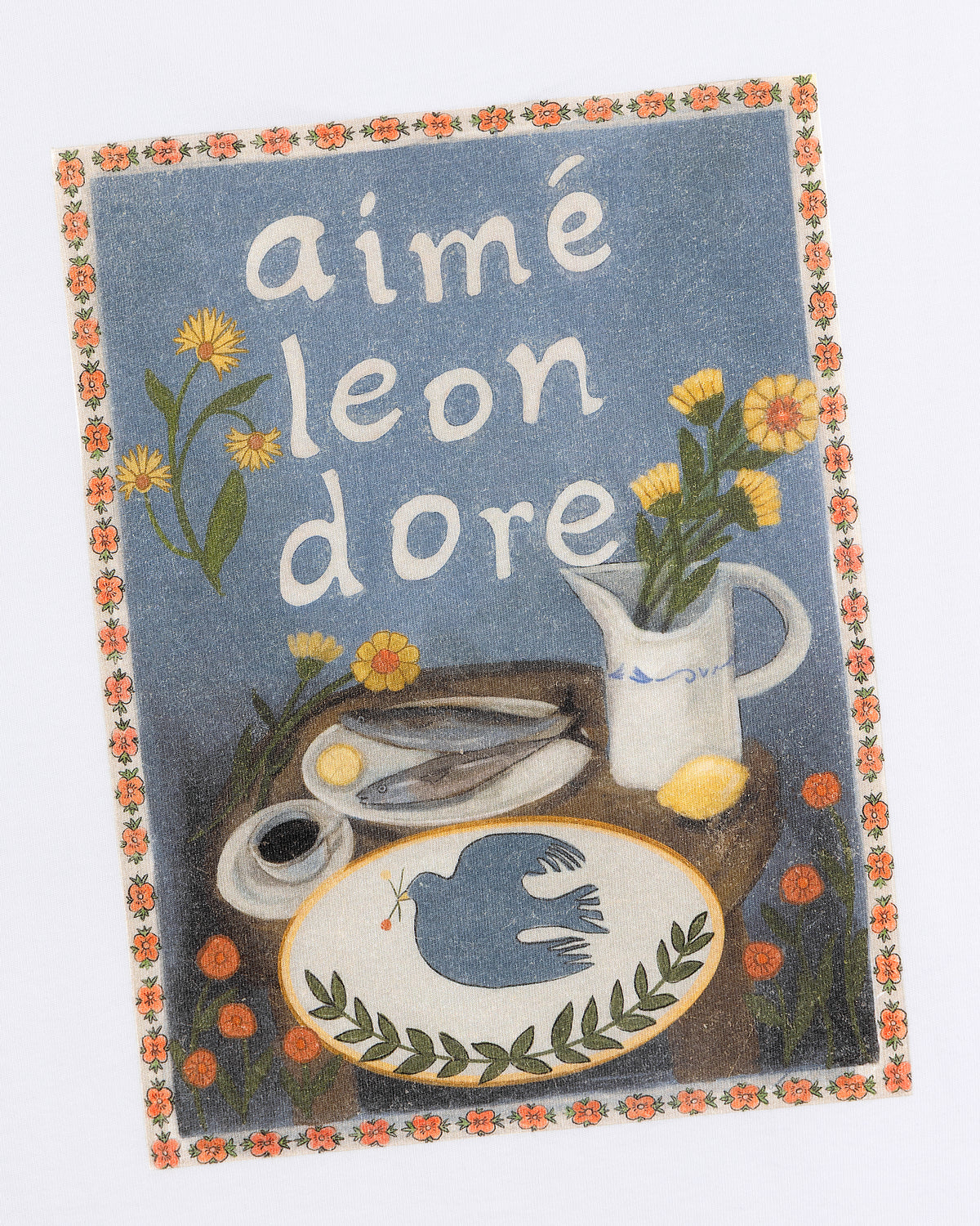 Breakfast Tee features a floral border with flowers, a bird, pitcher, scissors, lemon, and Aimé Leon Dore on a table.