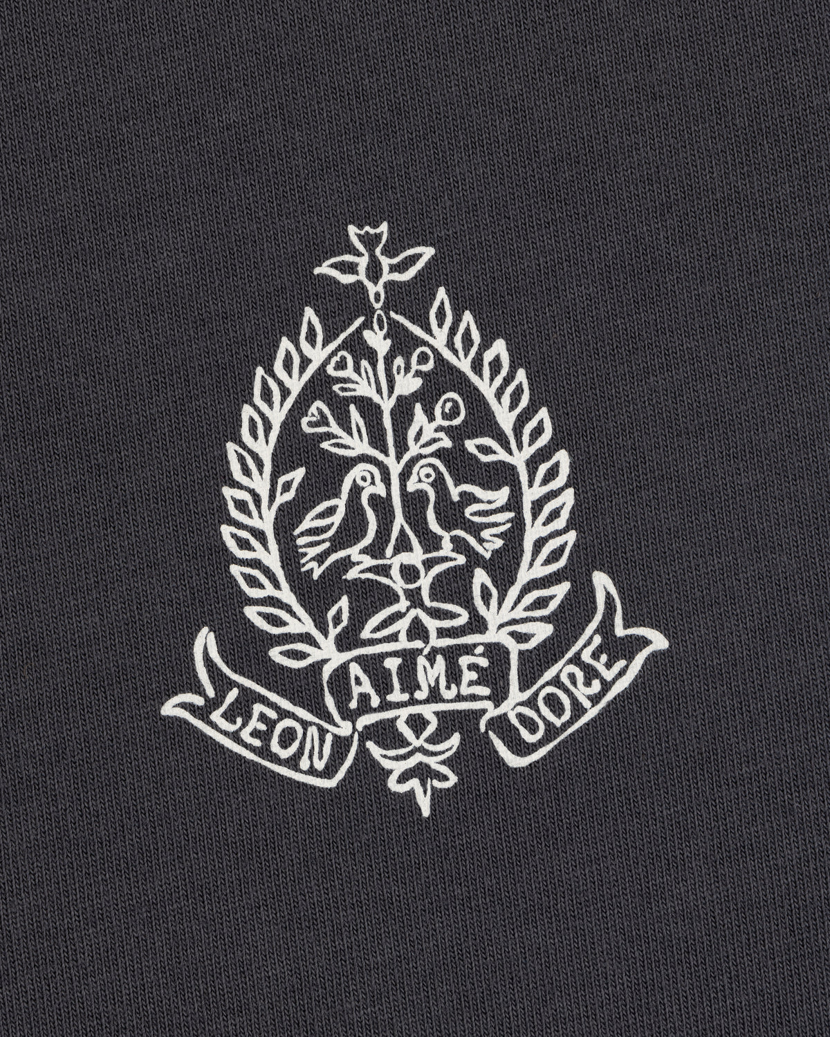 White embroidery of two birds, leafy branches, and "LEON AIME DOPE" on the Saint George Graphic Tee.