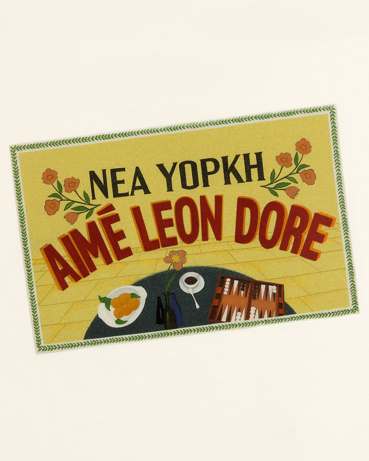 Rectangular patch on Aimé Postcard Tee with NEA YOPKH AIMÉ LEON DORE, flowers, food, cup, and books.