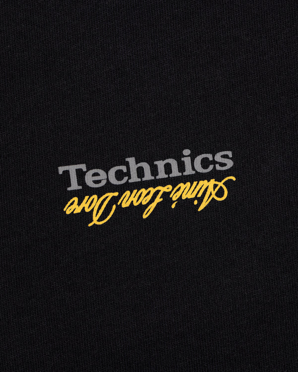 The ALD / Technics Logo Tee features gray "Technics" and yellow aline icon dore printed upside down on black fabric.