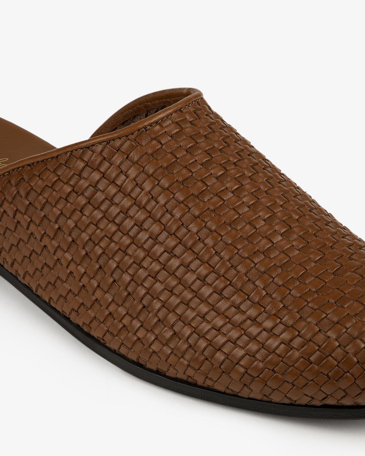 Close-up of the Leather Raffia Mule, a brown woven leather slip-on with a black sole on a white background.