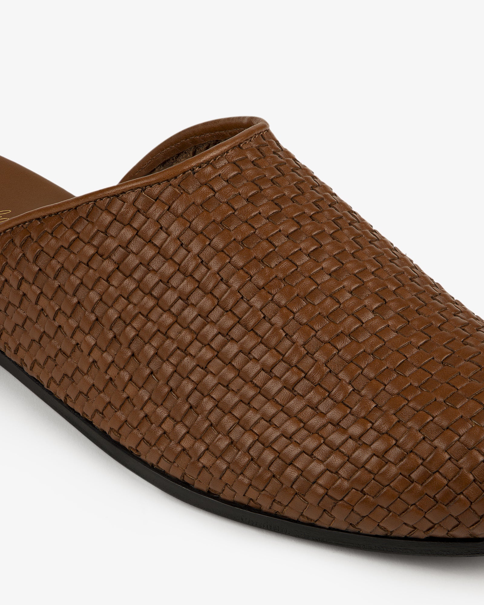 Close-up of the Leather Raffia Mule, a brown woven leather slip-on with a black sole on a white background.