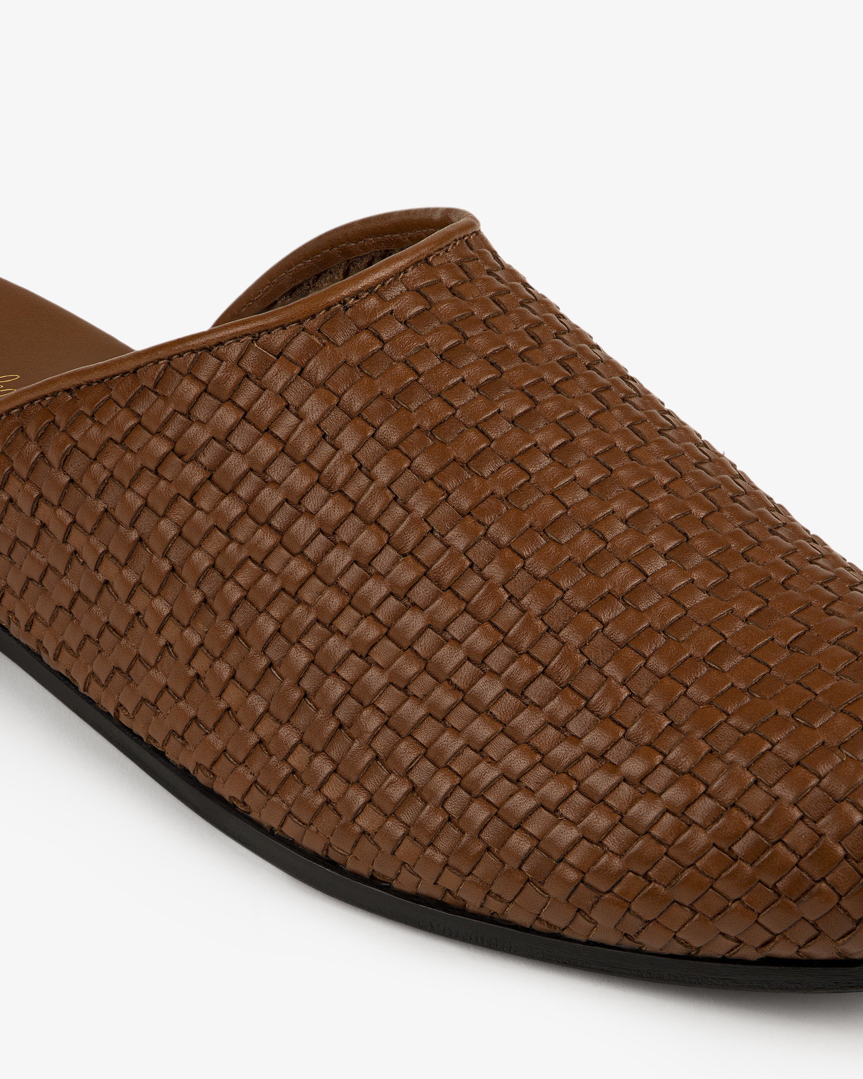 Close-up of the Leather Raffia Mule, a brown woven leather slip-on with a black sole on a white background.