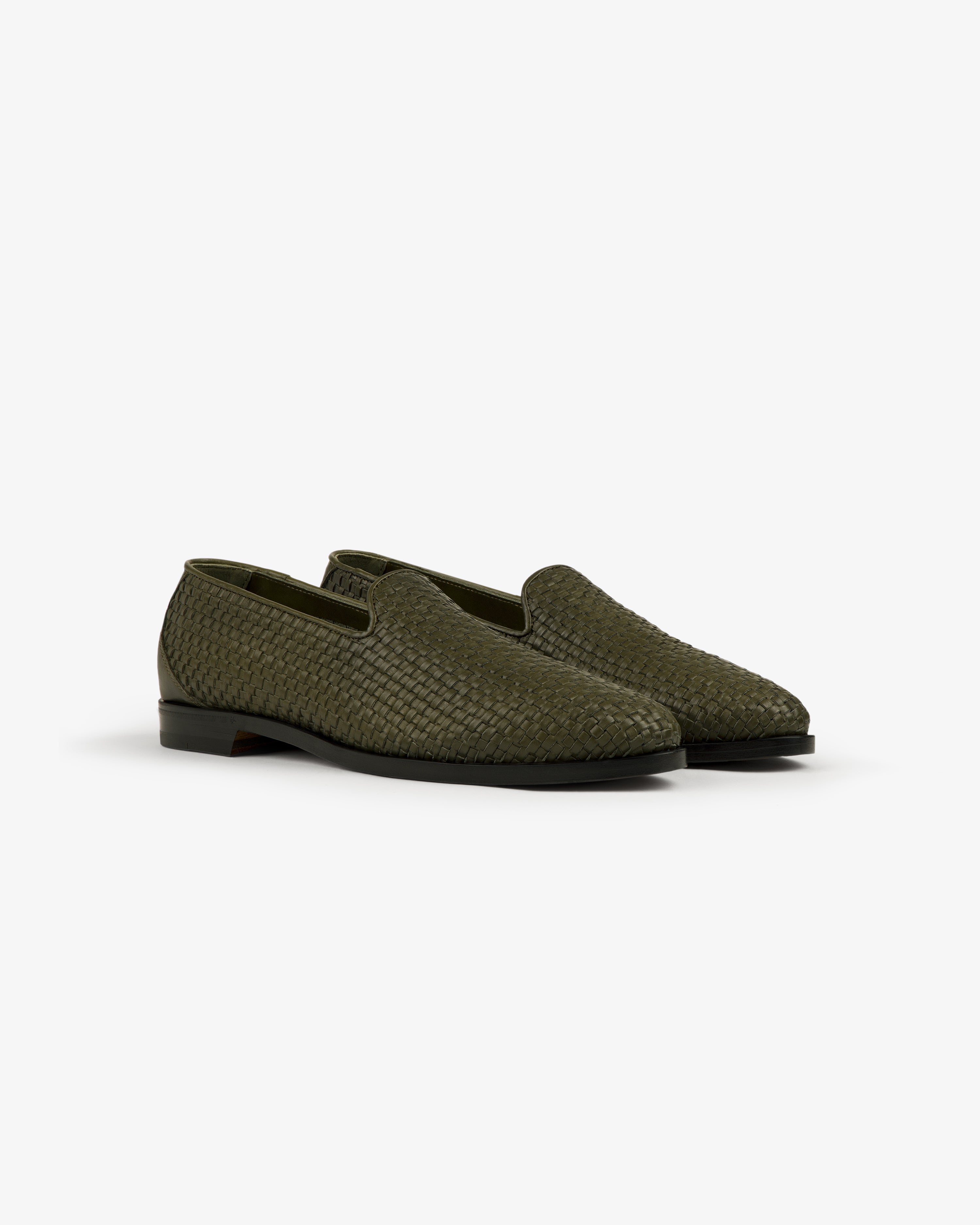 The Leather Raffia Loafer in green features black soles and a woven slip-on design, shown on a white background.