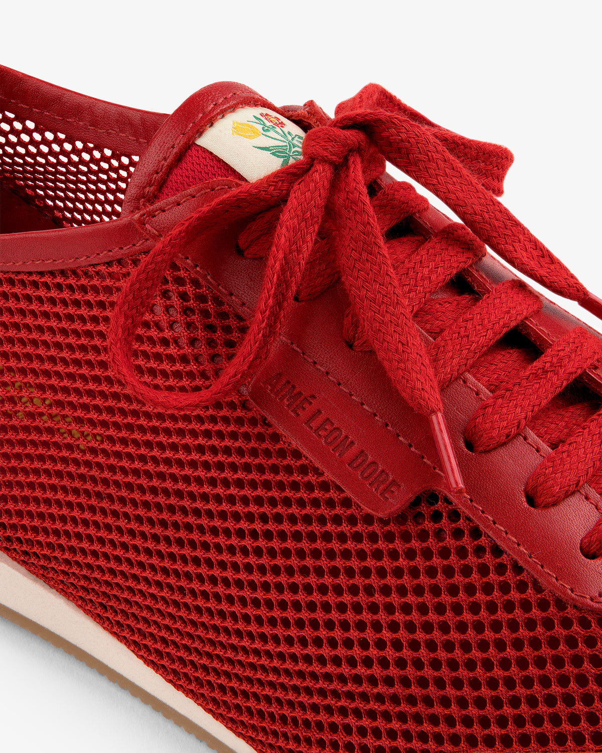 Close-up of a red Low Profile Mesh Trainer with red laces and AIME LEON DORE text on the lace holder.