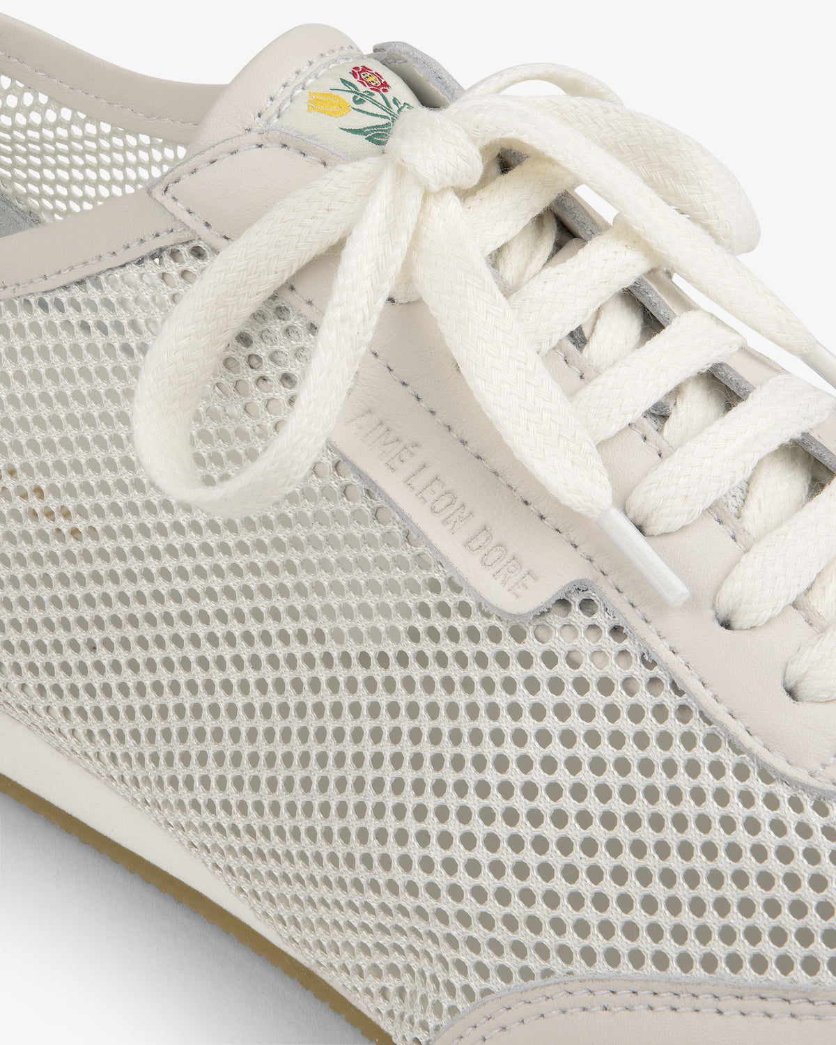 Close-up of the Low Profile Mesh Trainer, a white mesh sneaker with cream laces and a small flower on the tongue.