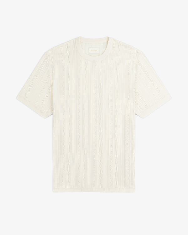 The Merino Wool Eyelet Tee is a short-sleeve, cream knit T-shirt with a subtle textured pattern, shown flat on white.