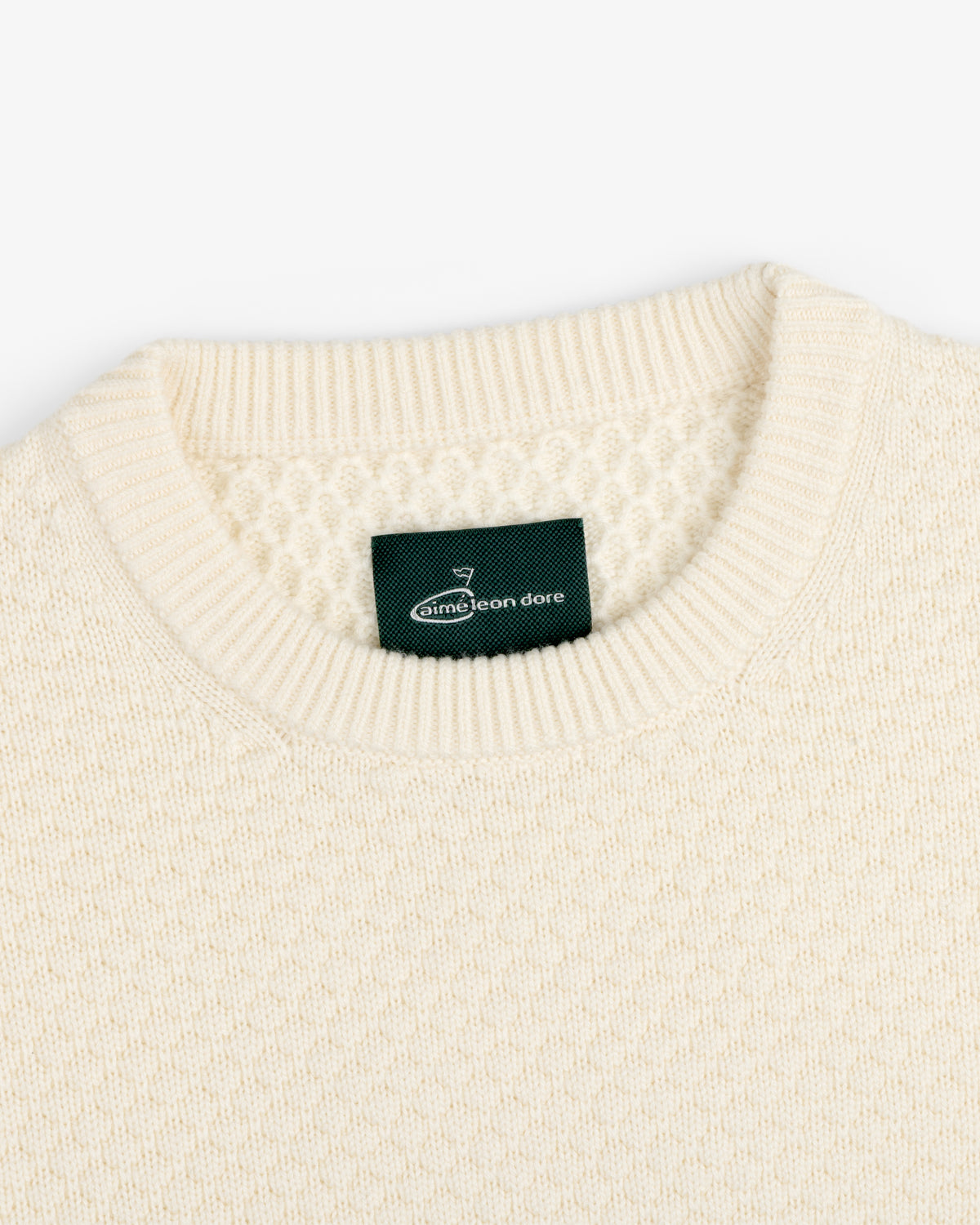 Close-up of the ALD Golf Clubhouse Sweater, a cream knit with a ribbed crew neck and green Aimé Leon Dore label.