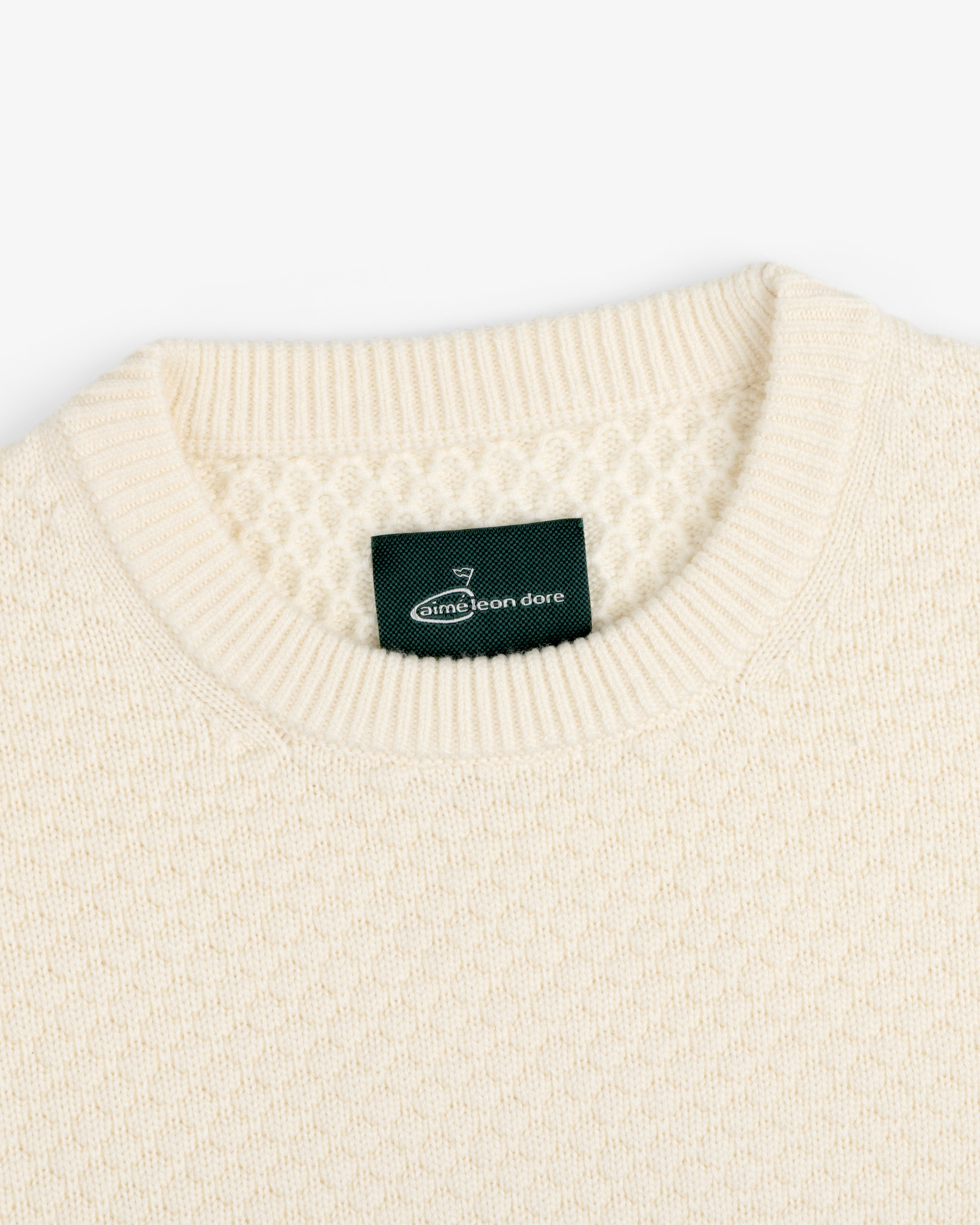Close-up of the ALD Golf Clubhouse Sweater, a cream knit with a ribbed crew neck and green Aimé Leon Dore label.