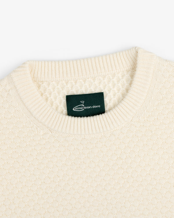 Close-up of the ALD Golf Clubhouse Sweater, a cream knit with a ribbed crew neck and green Aimé Leon Dore label.
