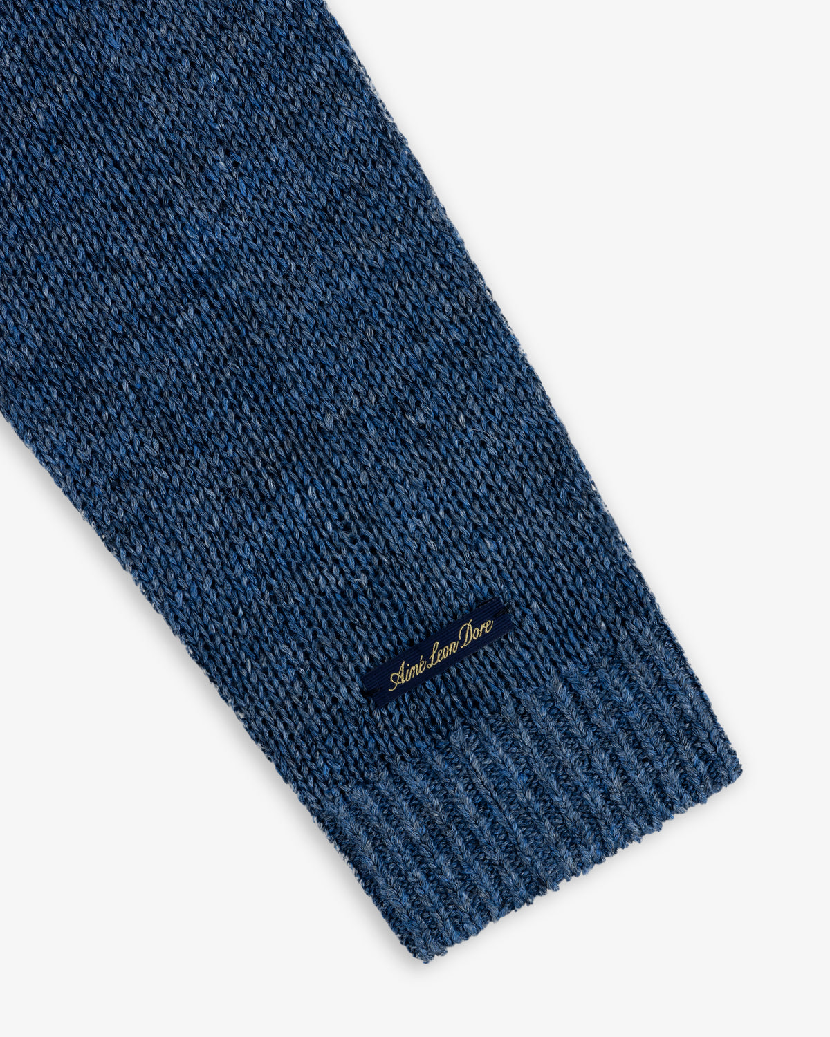 Close-up of a blue Linen Knit Crewneck Sweater sleeve with a small black Aimé Leon Dore label in gold script.