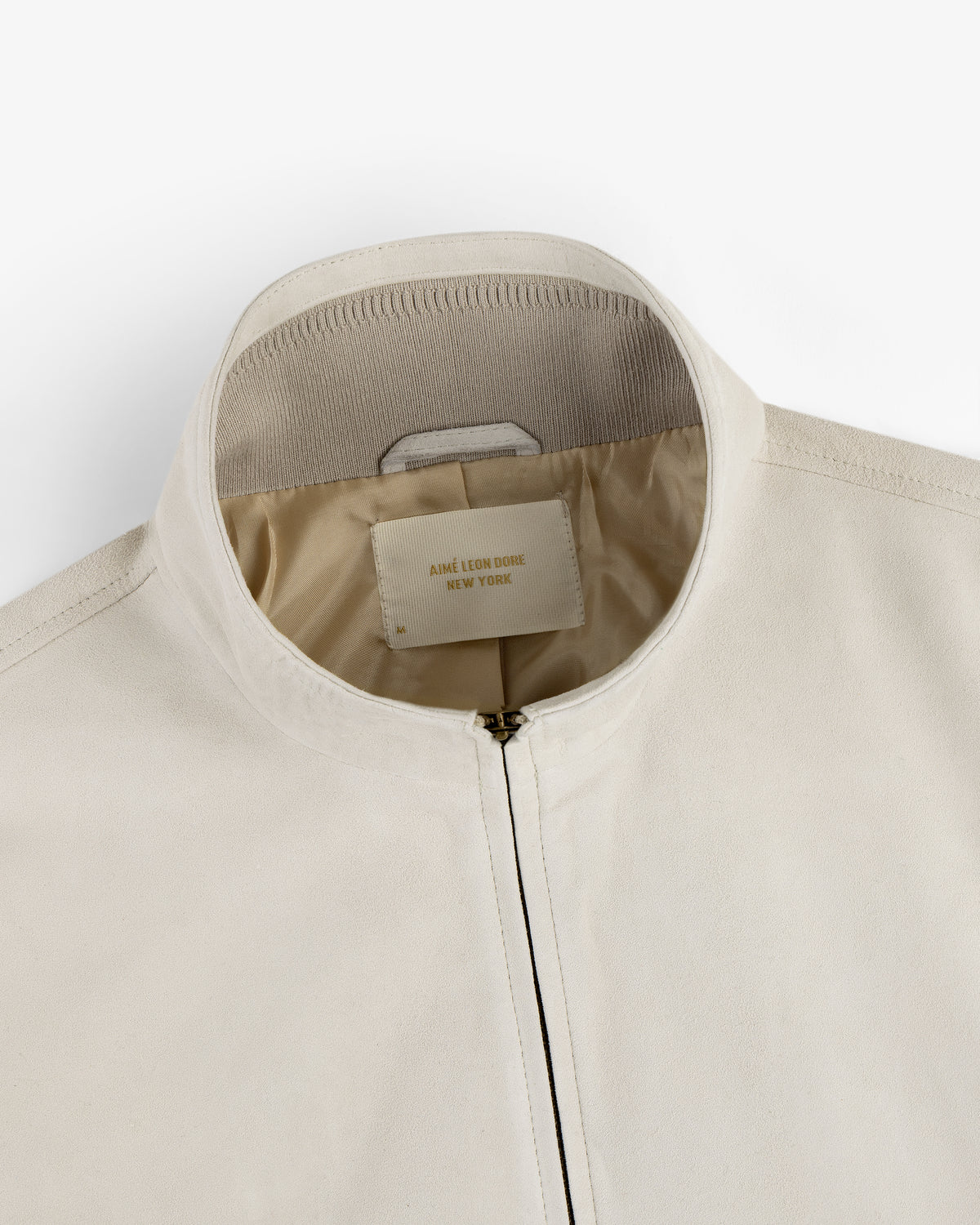 Close-up of a cream suede track jacket with zipper and a JANE LÉON DORE NEW YORK label inside the collar.