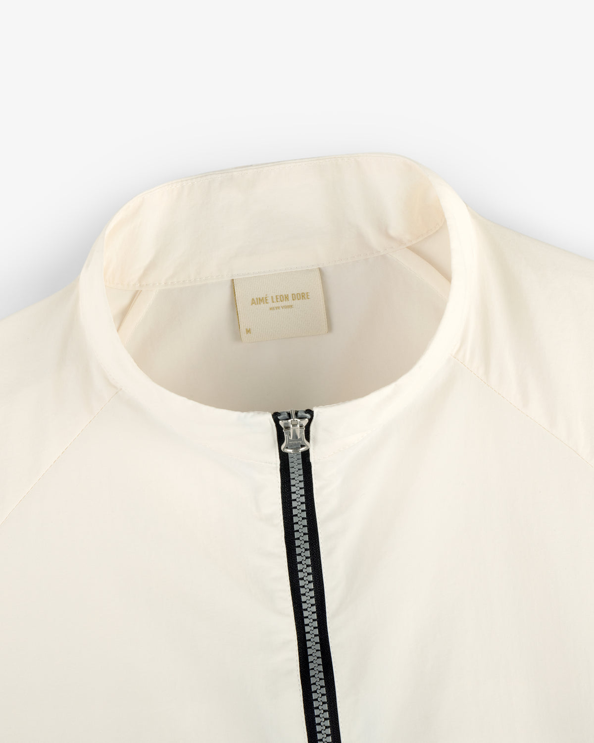 Close-up of a white Nylon Packable Windbreaker with a zipper and Aimé Leon Dore label inside the collar.