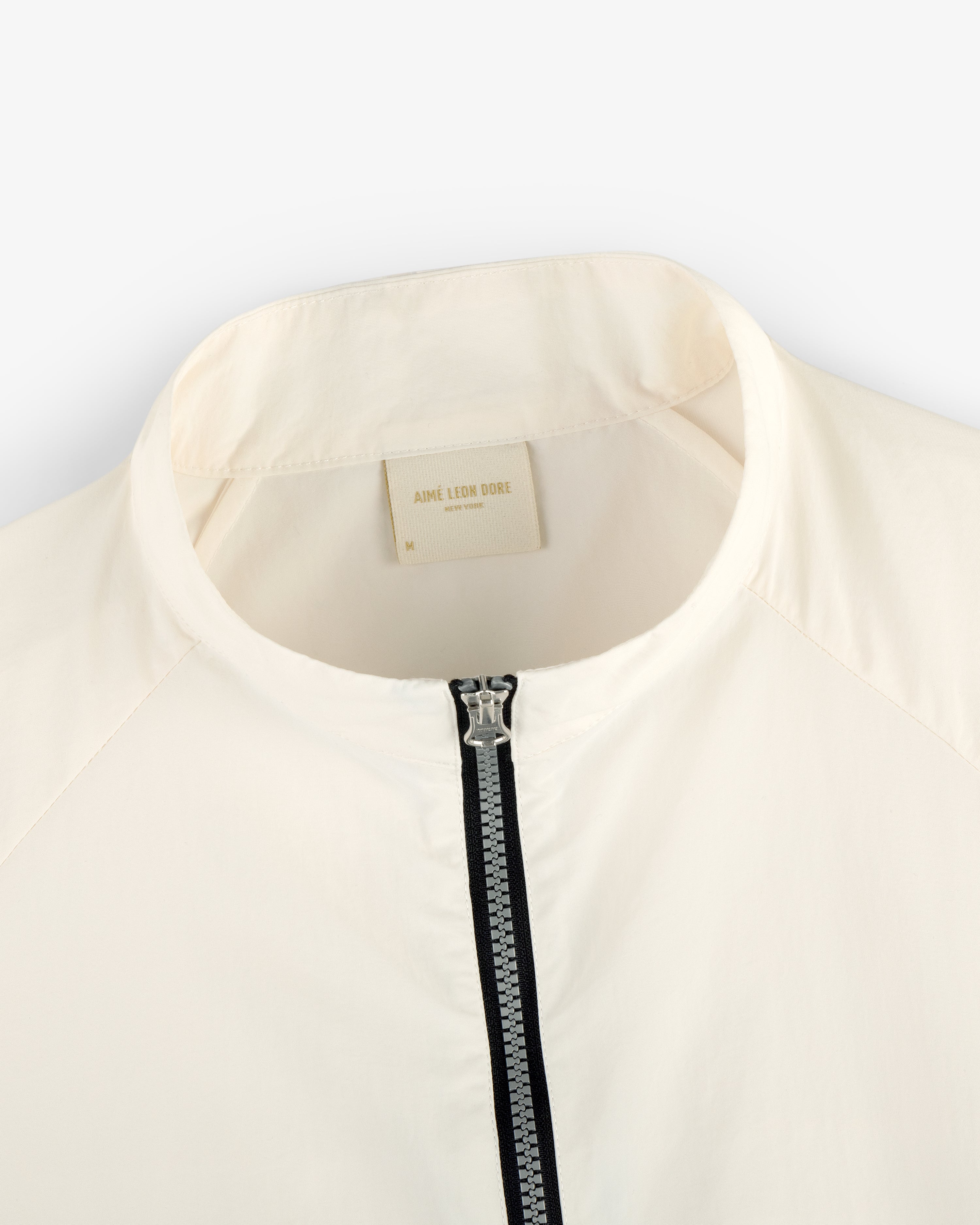Close-up of a white Nylon Packable Windbreaker with a zipper and Aimé Leon Dore label inside the collar.
