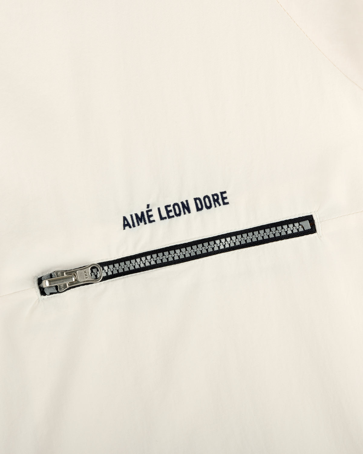 Close-up of an off-white Nylon Packable Windbreaker with a black zipper and AIMÉ LEON DORE text above the zipper.